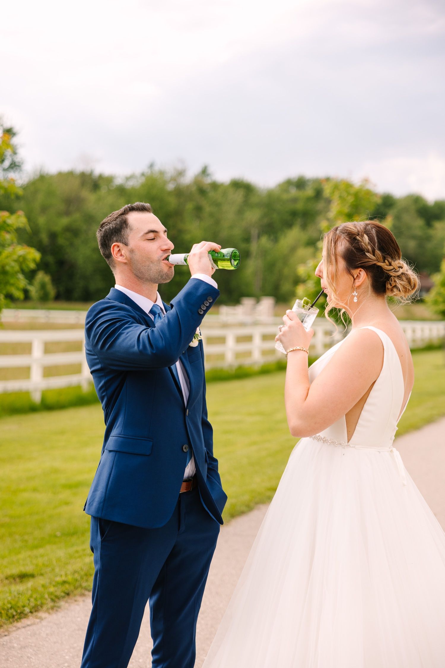 A couple in wedding attire drinks from bottles during their outdoor countryside photo session.