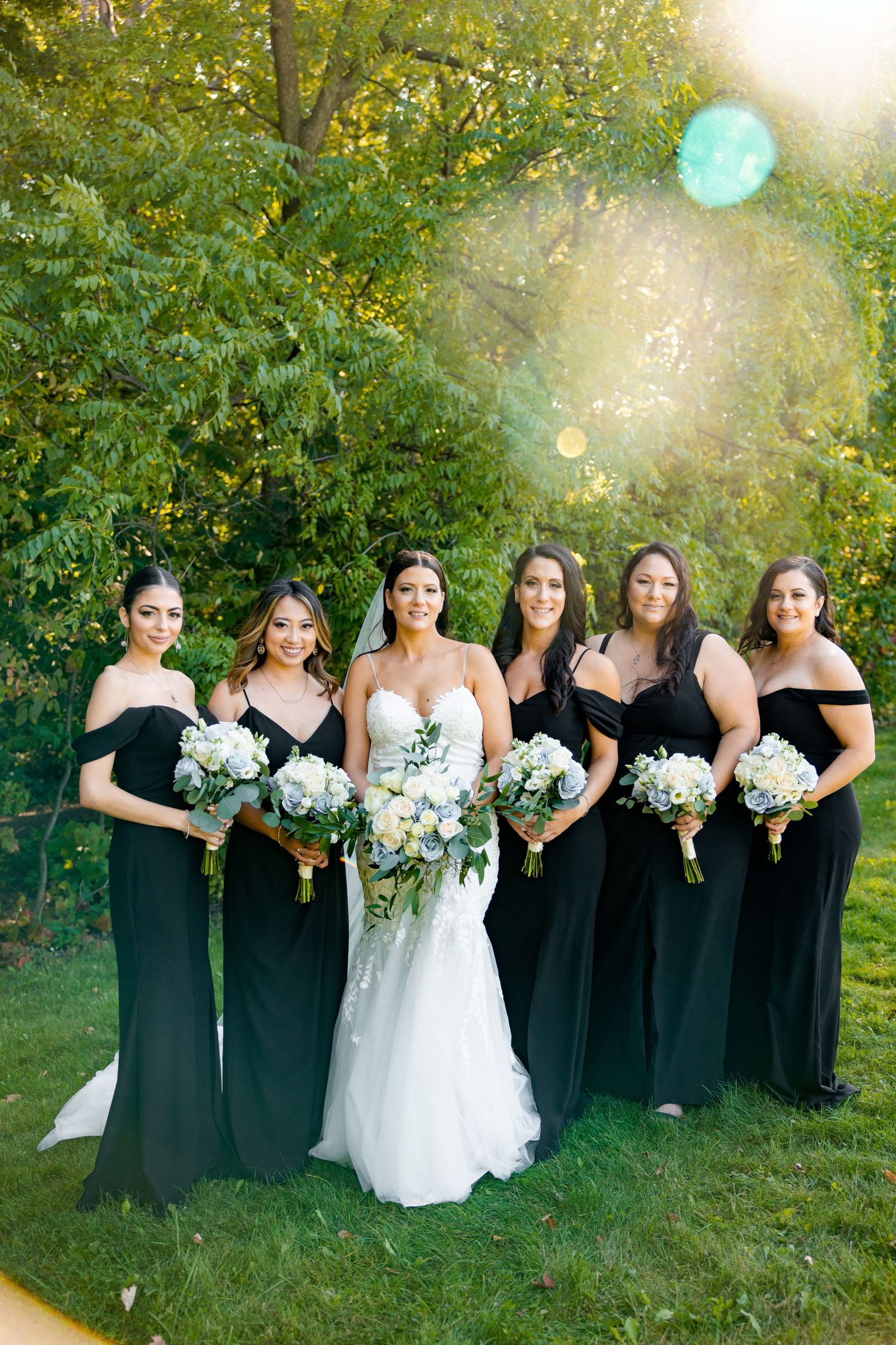 Bridesmaids in elegant black dresses hold white bouquets while standing with bride in white gown outdoors.