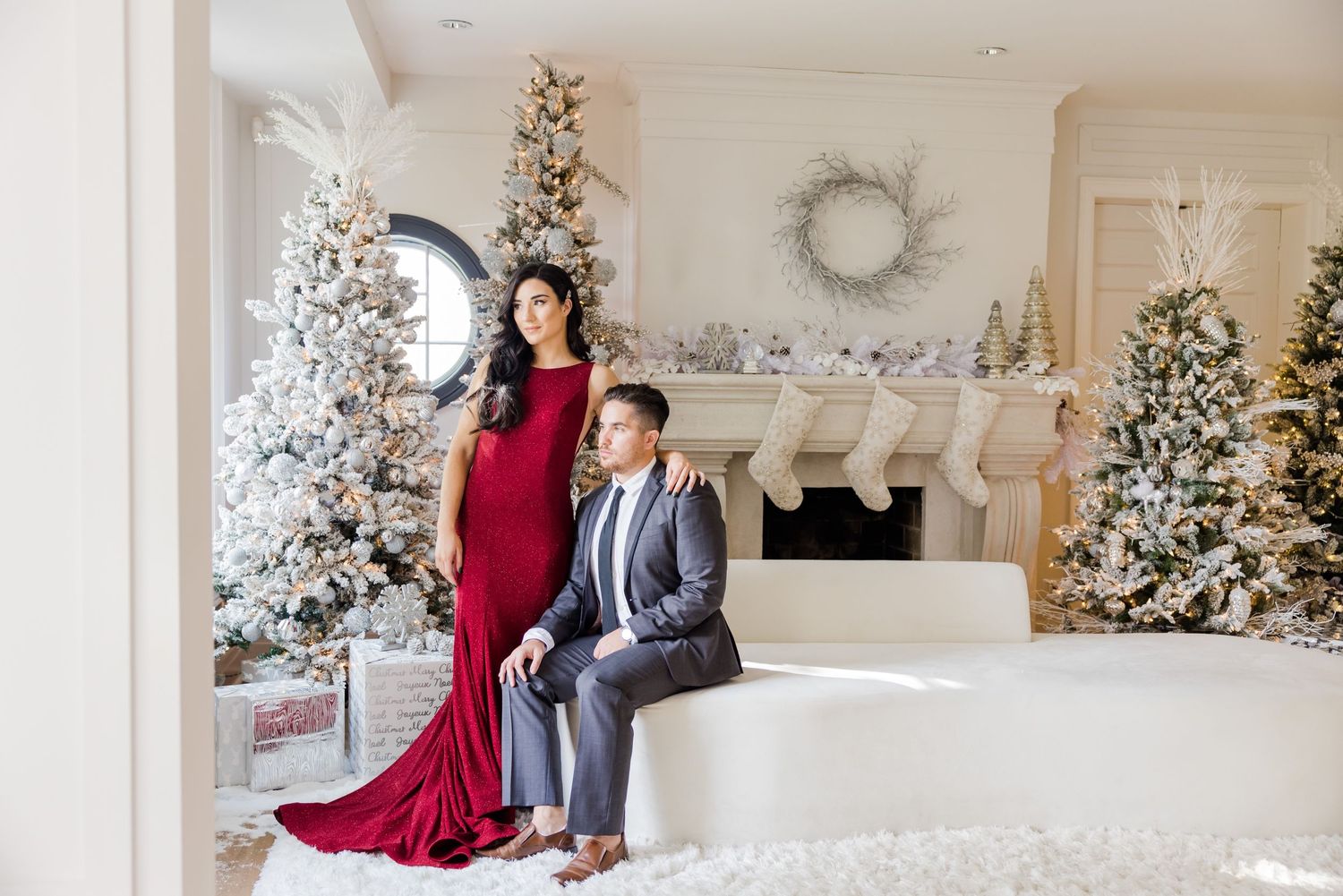 Elegant formal portrait with red gown against white Christmas trees and fireplace setting.