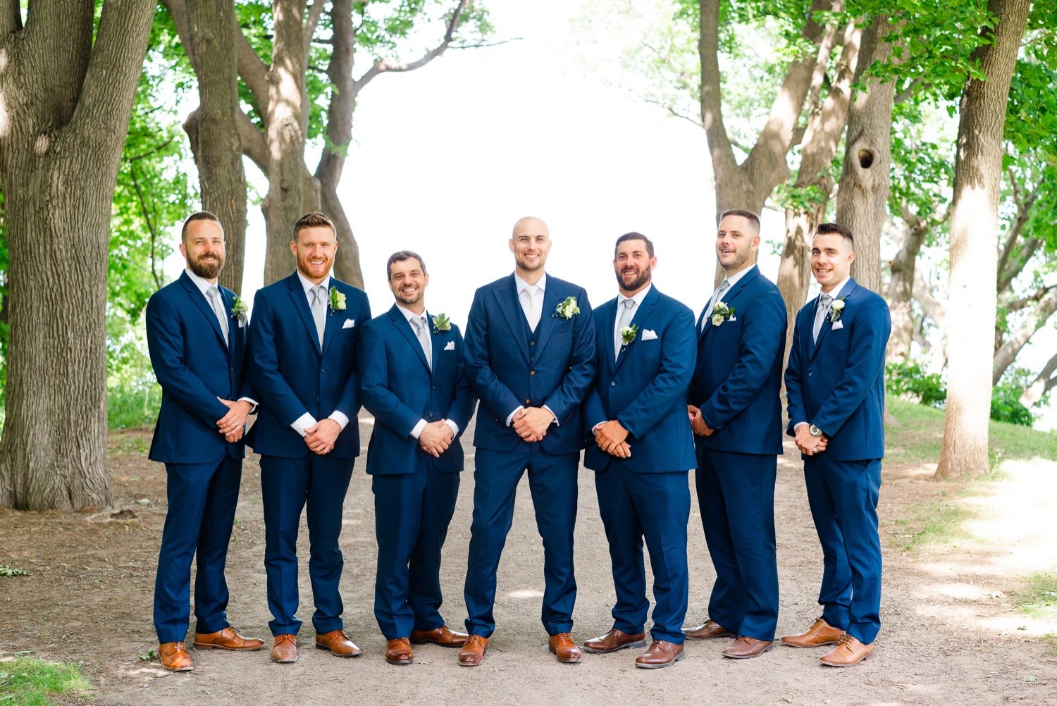 Wedding party of groomsmen in coordinated navy suits and brown shoes pose under green trees.