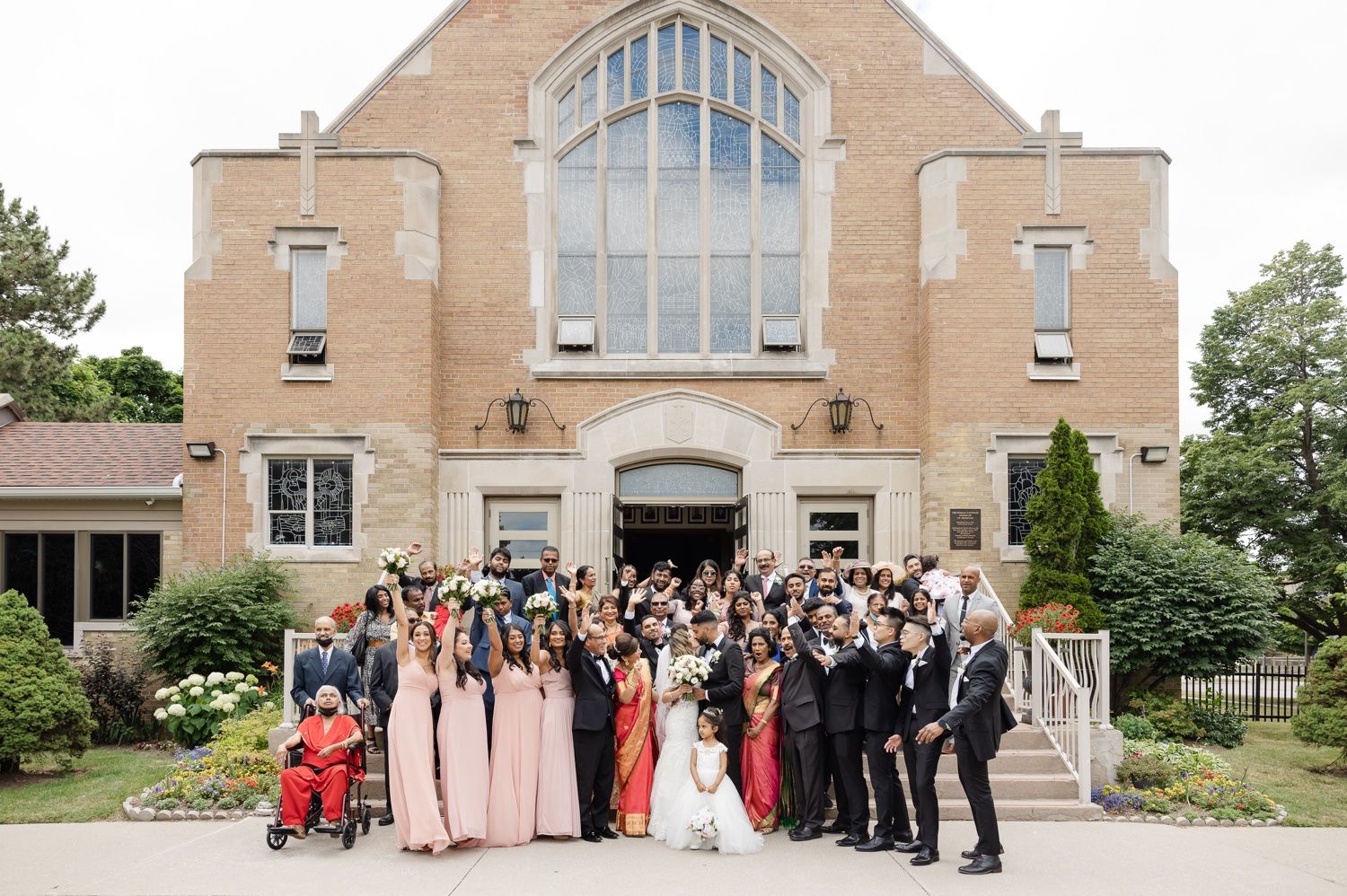 Group photo of wedding party and guests in front of tan brick church.