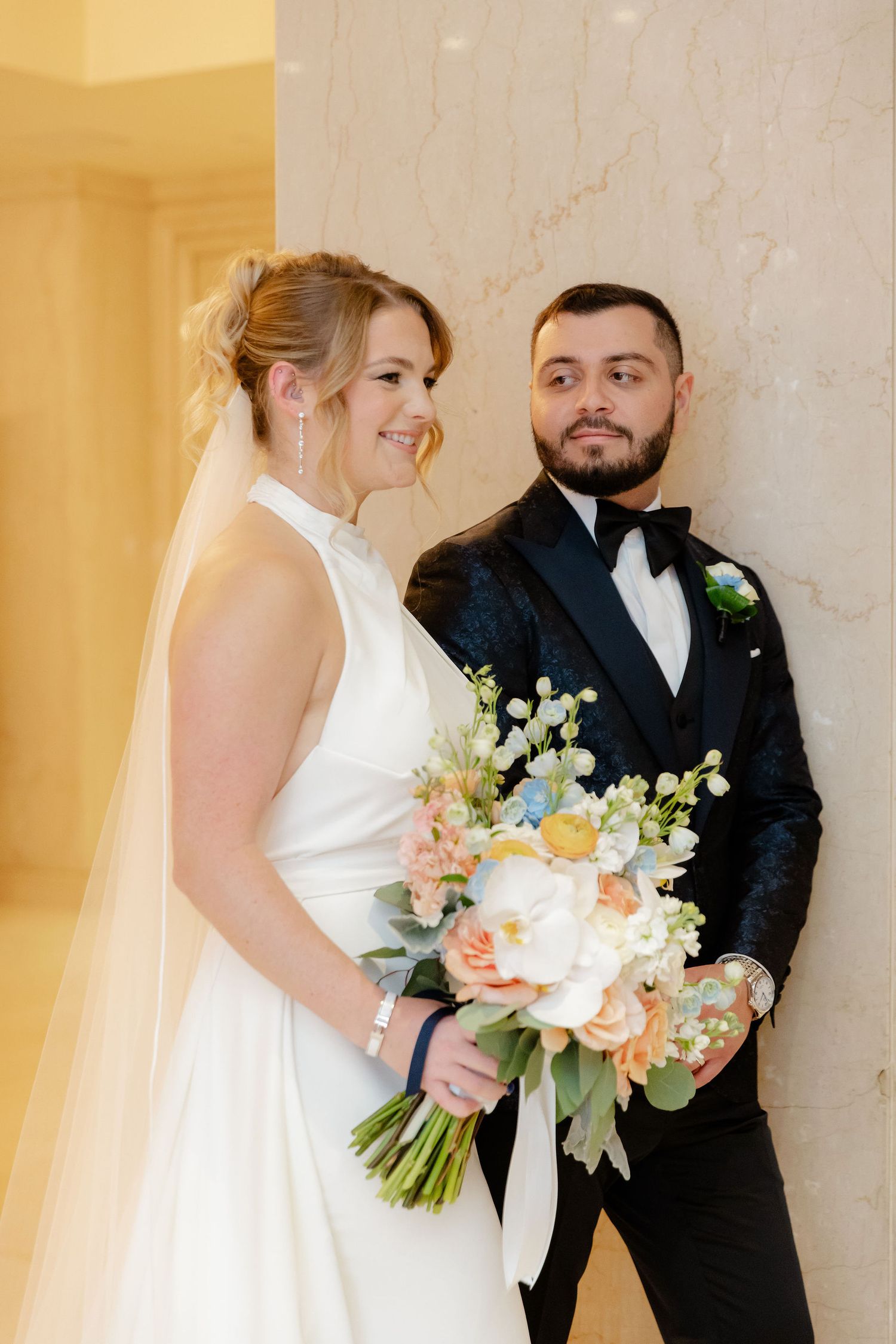 Bride in white gown and groom in black tuxedo share tender moments against marble wall.