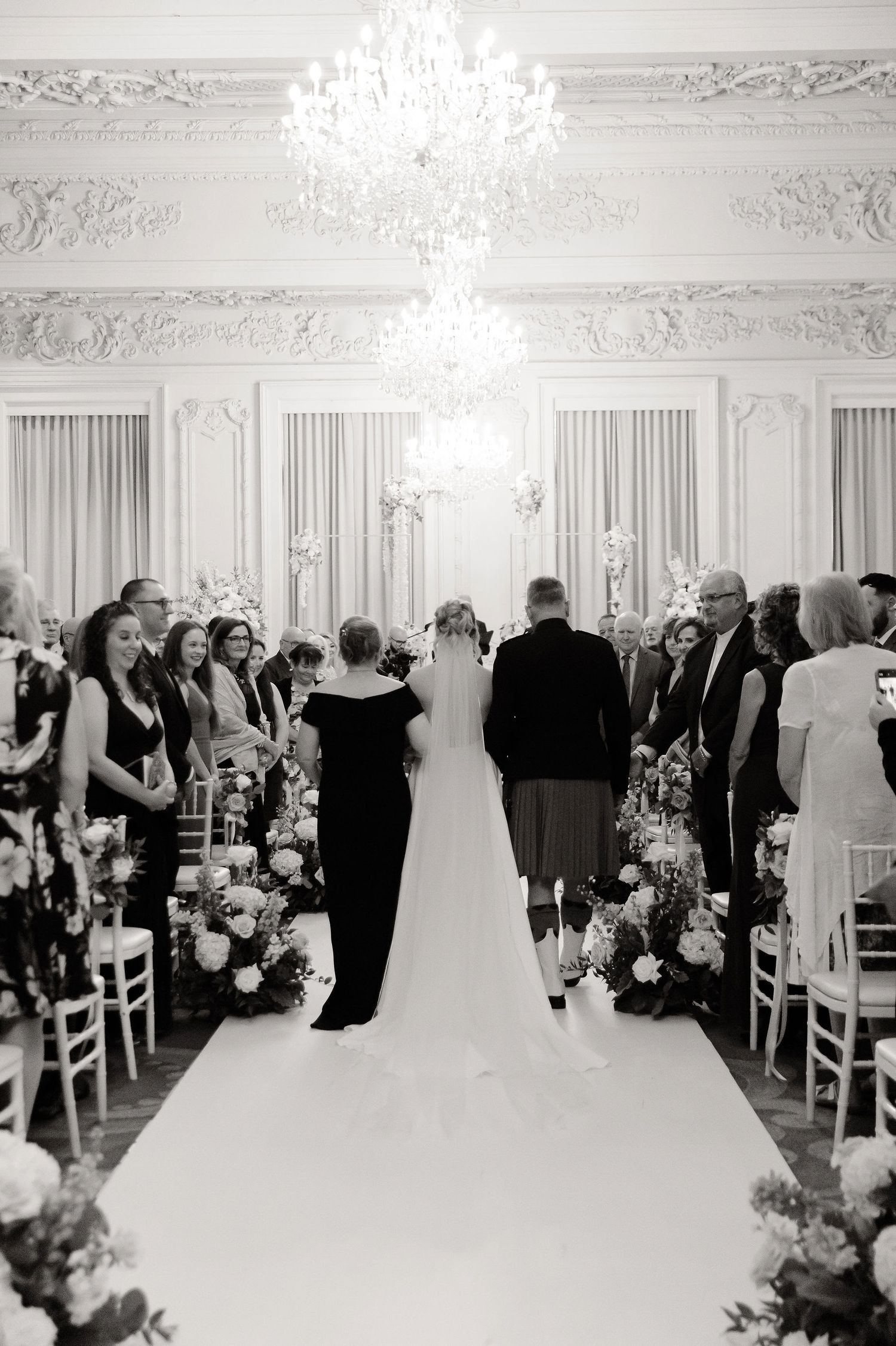 Wedding guests standing during ceremony in elegant white ballroom with chandelier.