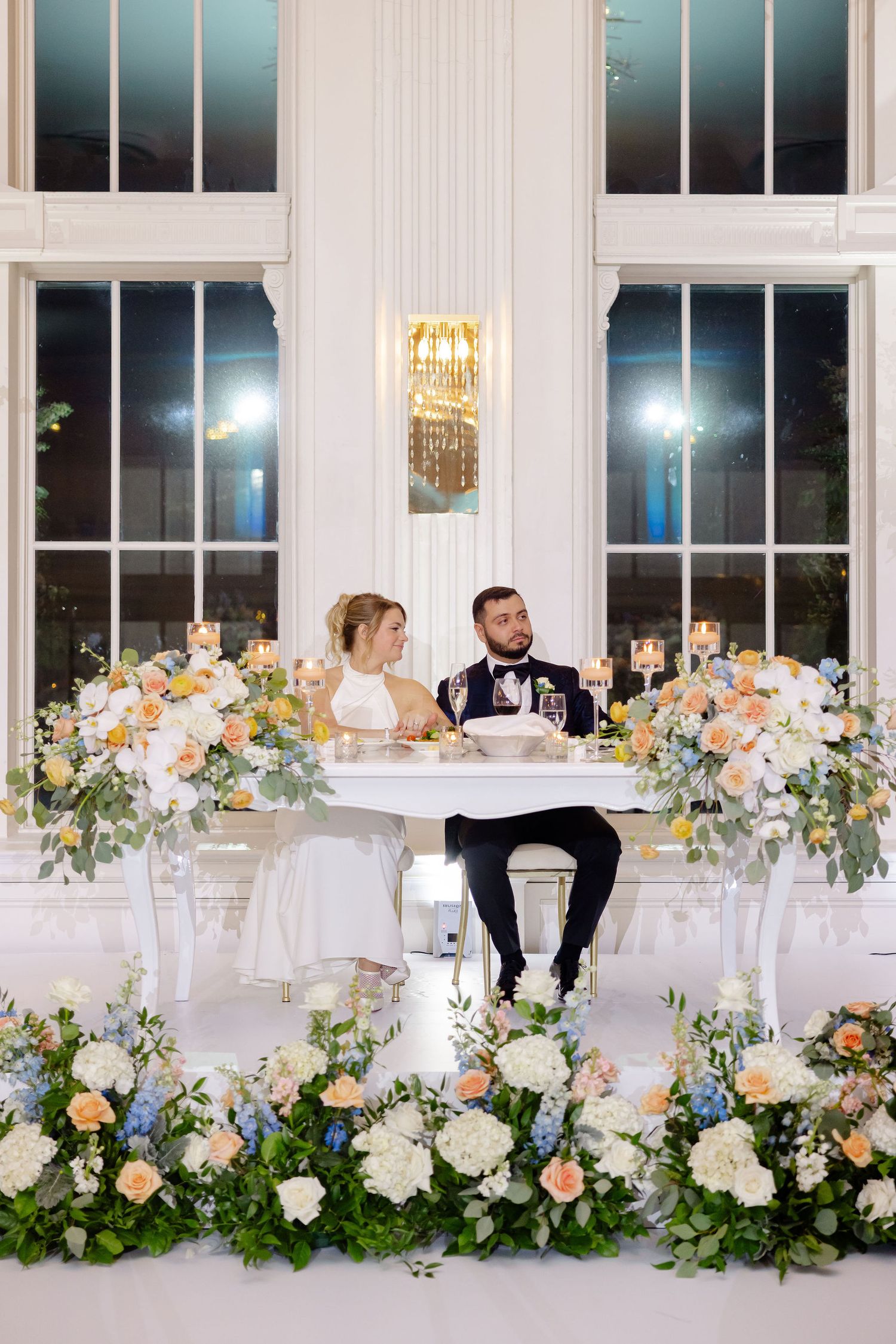 Wedding reception table adorned with colorful floral arrangements and crystal chandeliers in a bright white room.