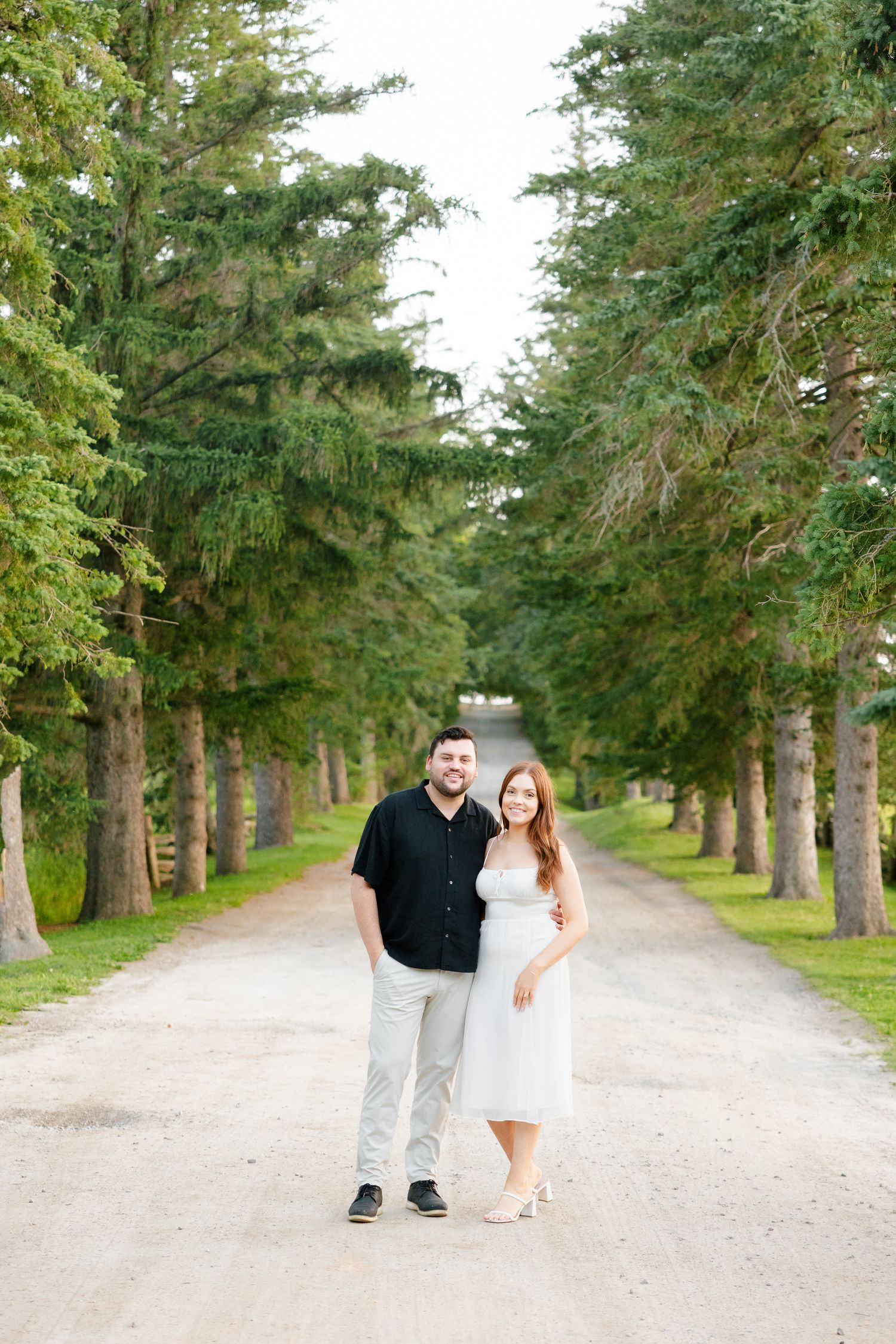 A couple stands together on a tree-lined path with evergreen trees creating a natural tunnel.