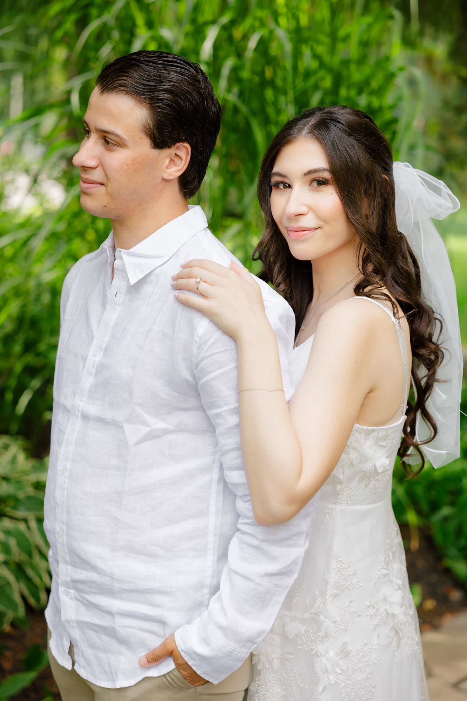 A romantic garden portrait of newlyweds in white attire against lush green foliage.