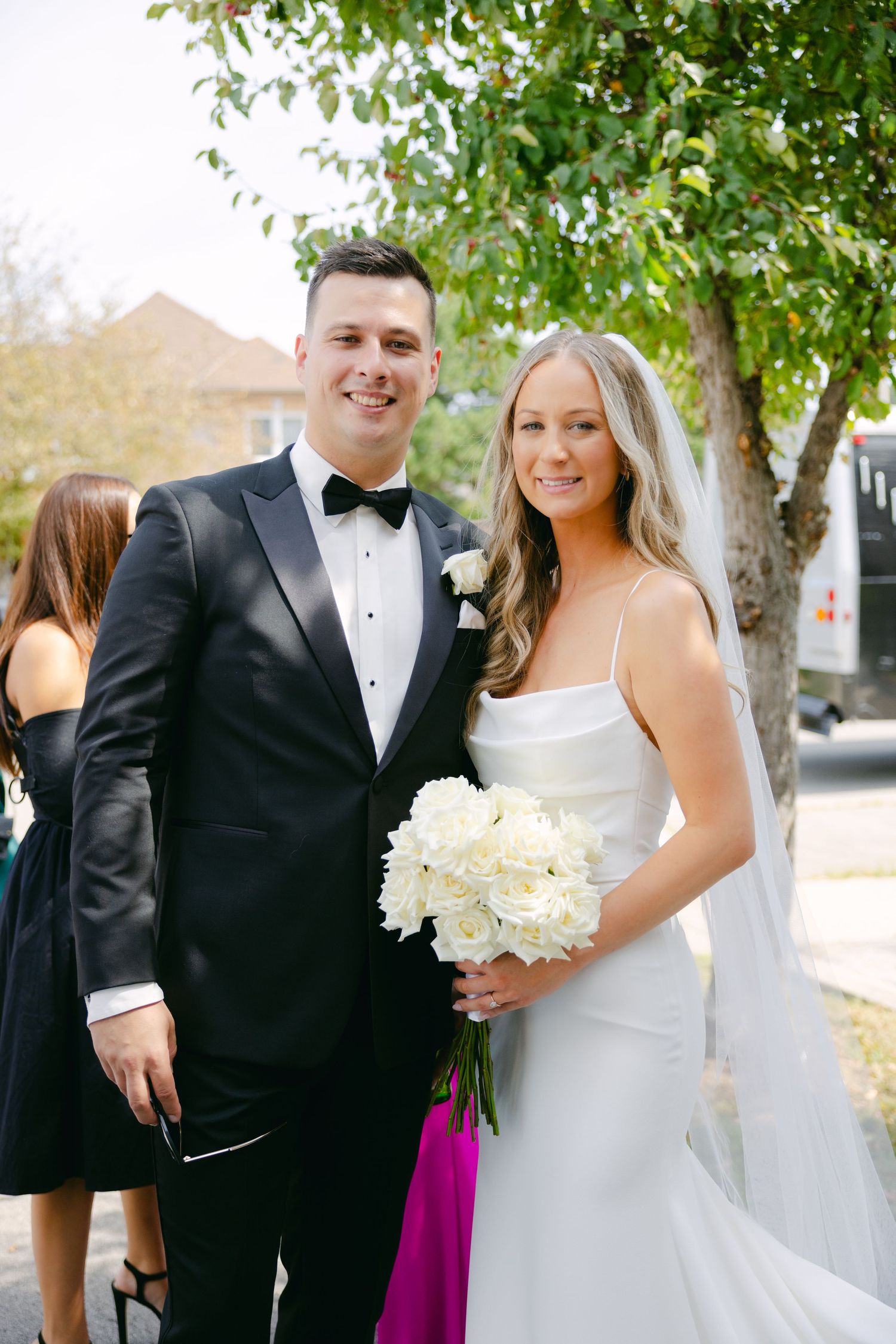 Elegant wedding couple in formal attire poses with white bouquet outdoors.
