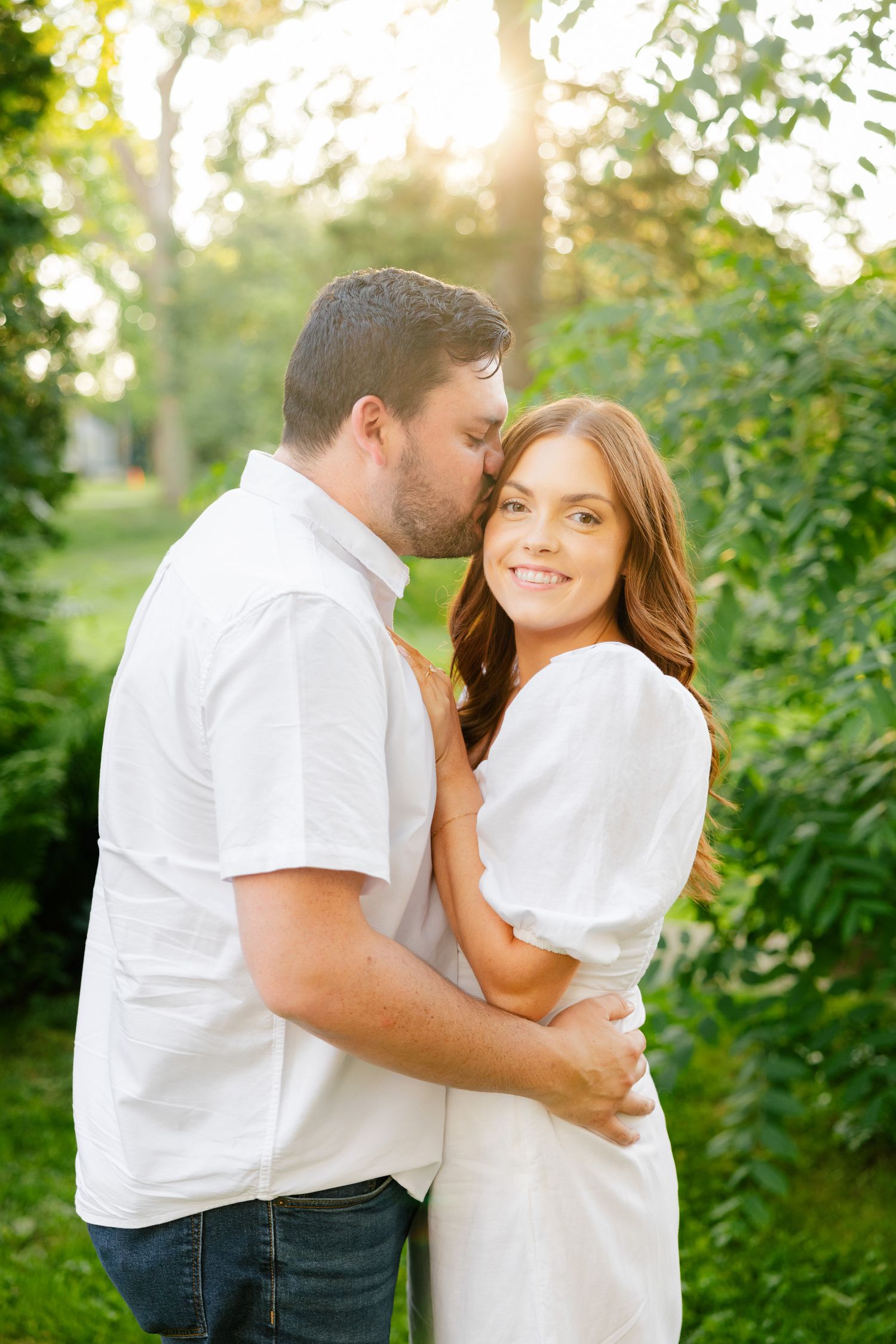 Romantic couple embracing in white outfits against sunlit garden background.