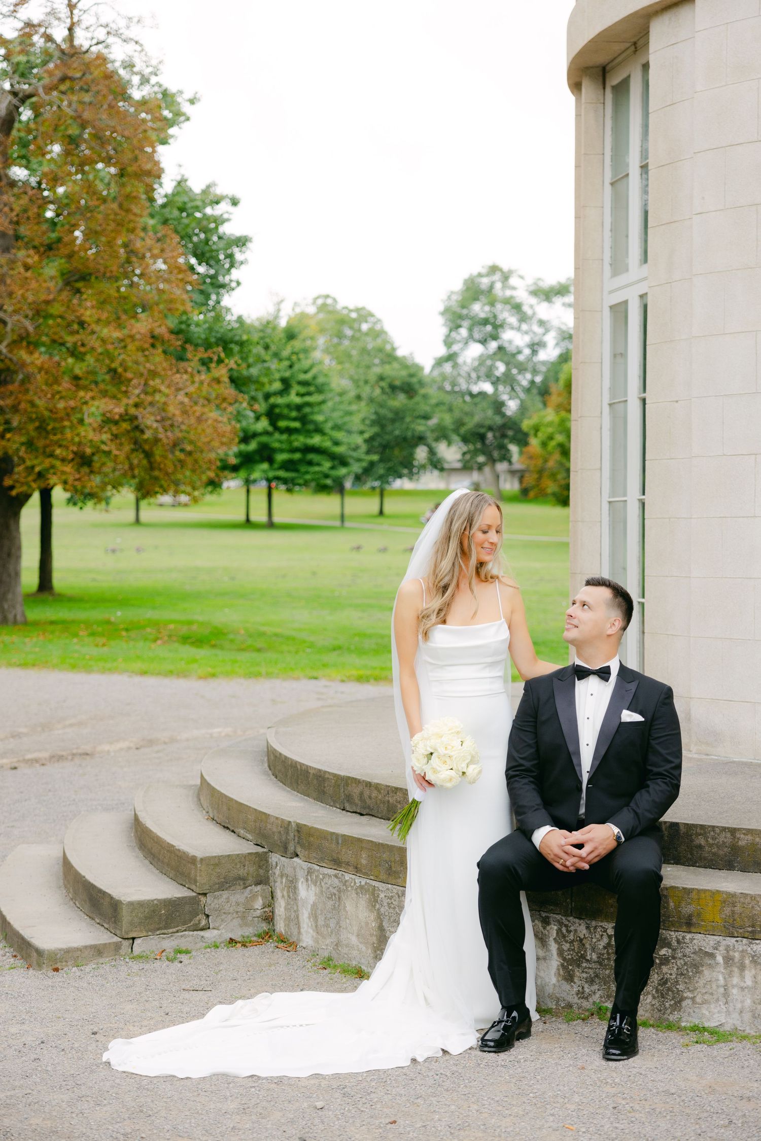 Wedding couple poses on stone steps with autumn trees in background.