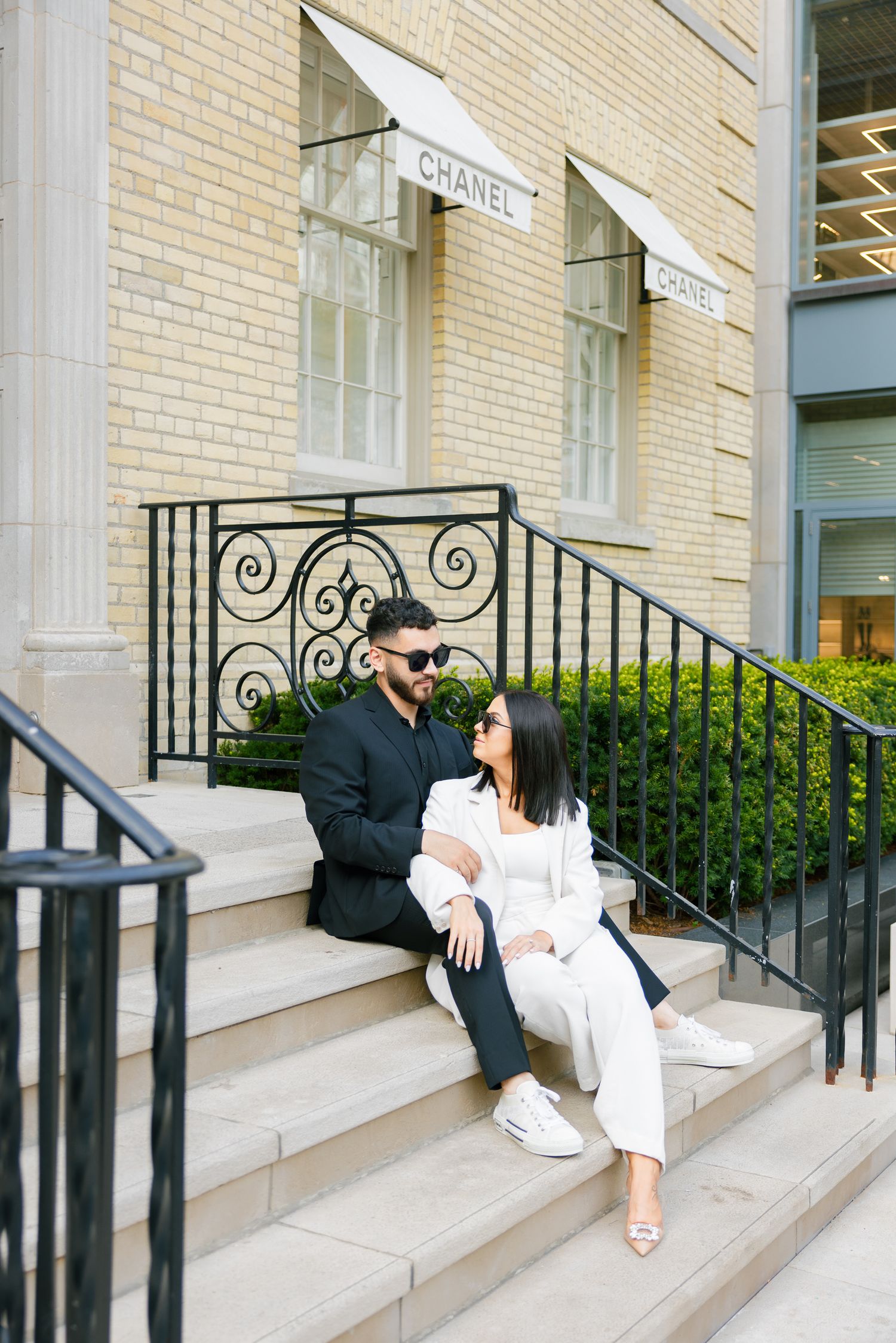 A couple in contrasting black and white outfits sit romantically on stone steps outside a Chanel boutique.