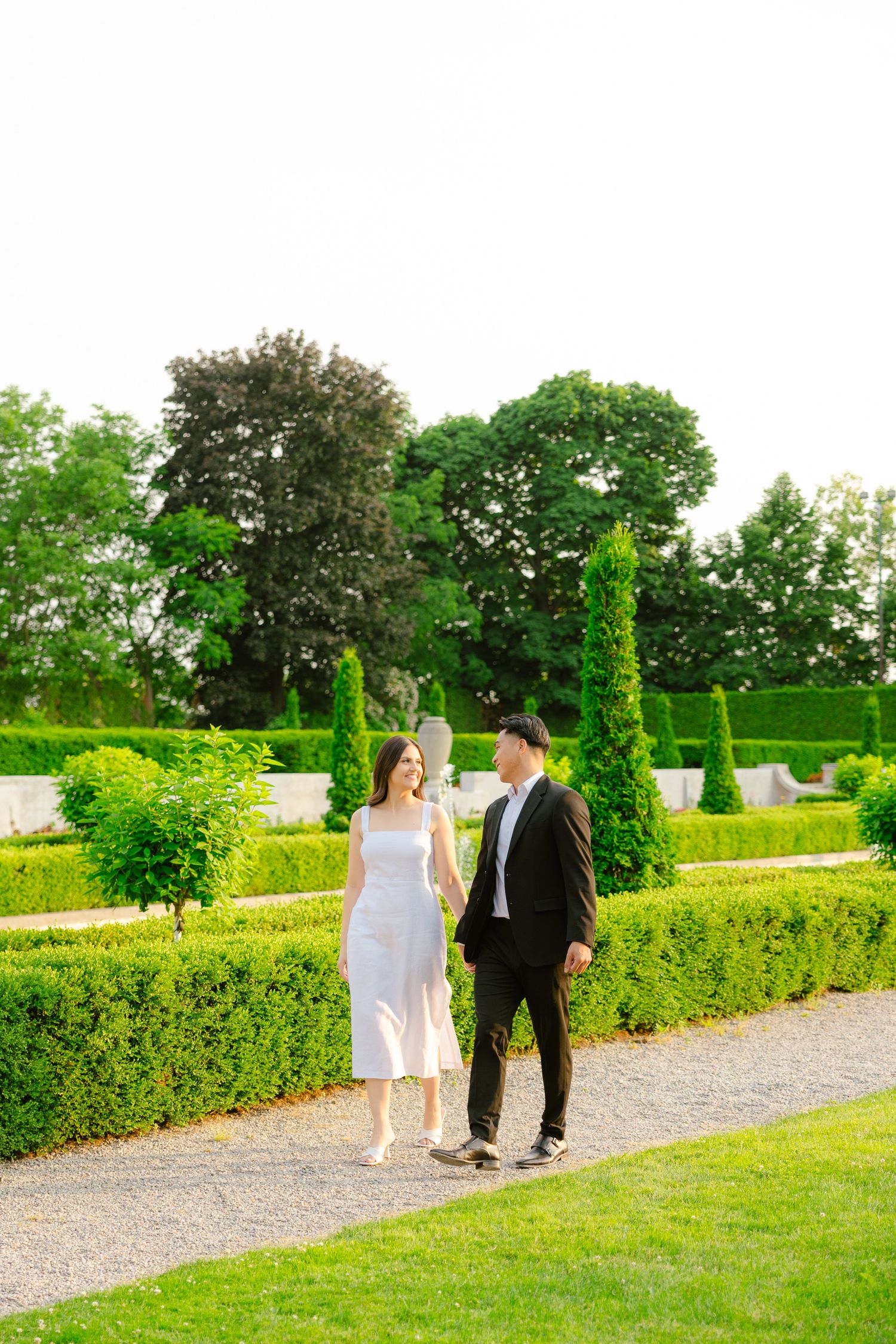 Elegantly dressed couple strolling along a gravel pathway in a formal garden with perfectly trimmed hedges and topiaries.