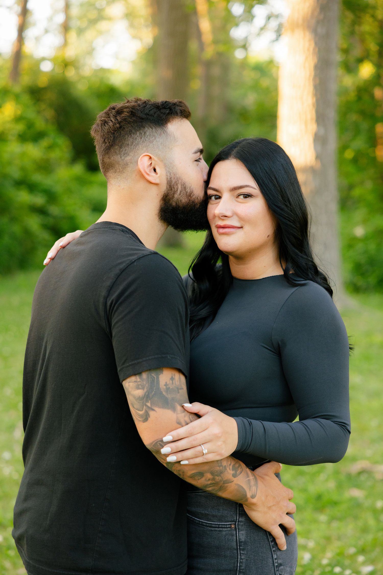 Affectionate couple in dark clothing embracing outdoors with golden sunlight filtering through trees.