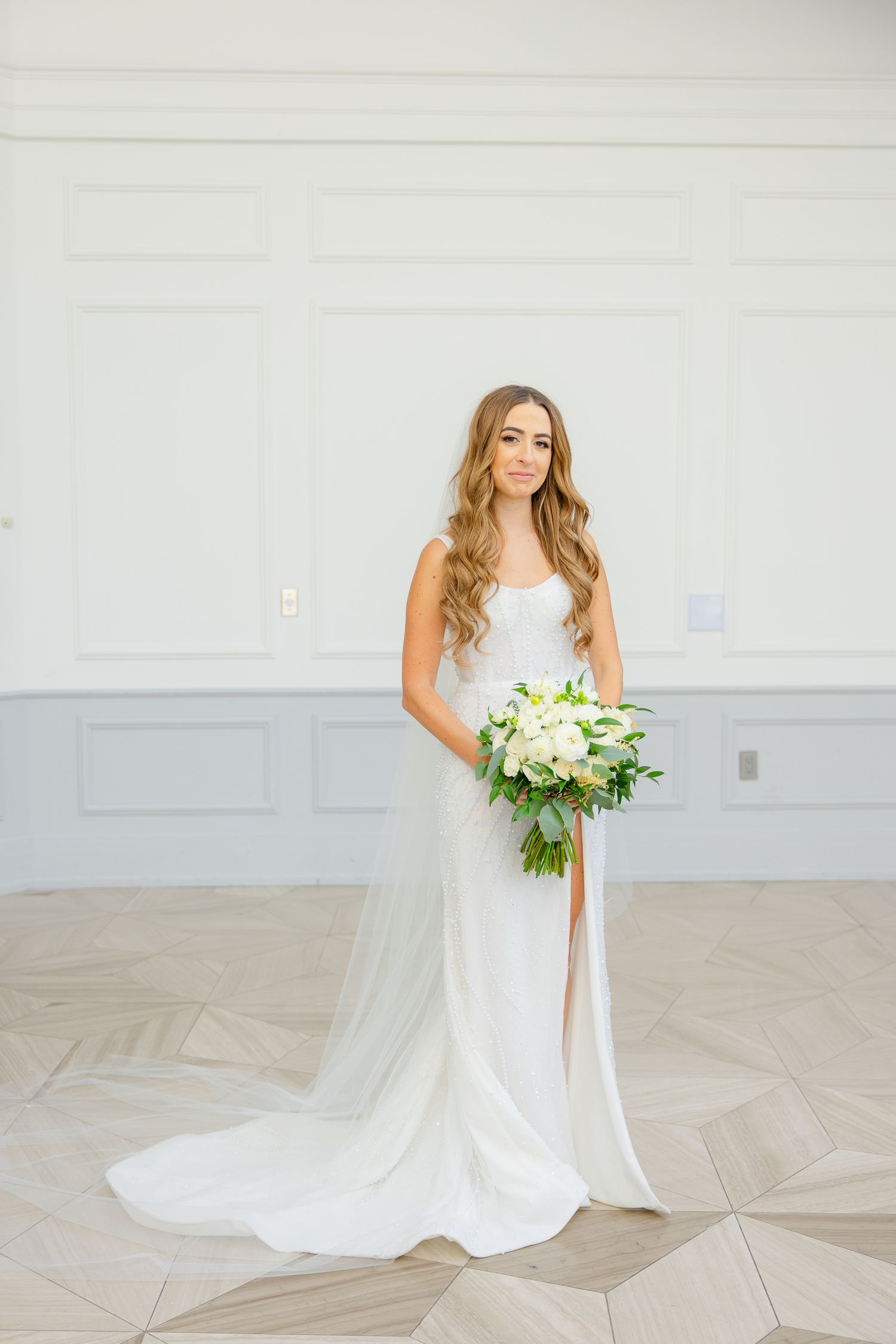 Bride in sleek white gown holds beautiful white and green bouquet against neutral backdrop.