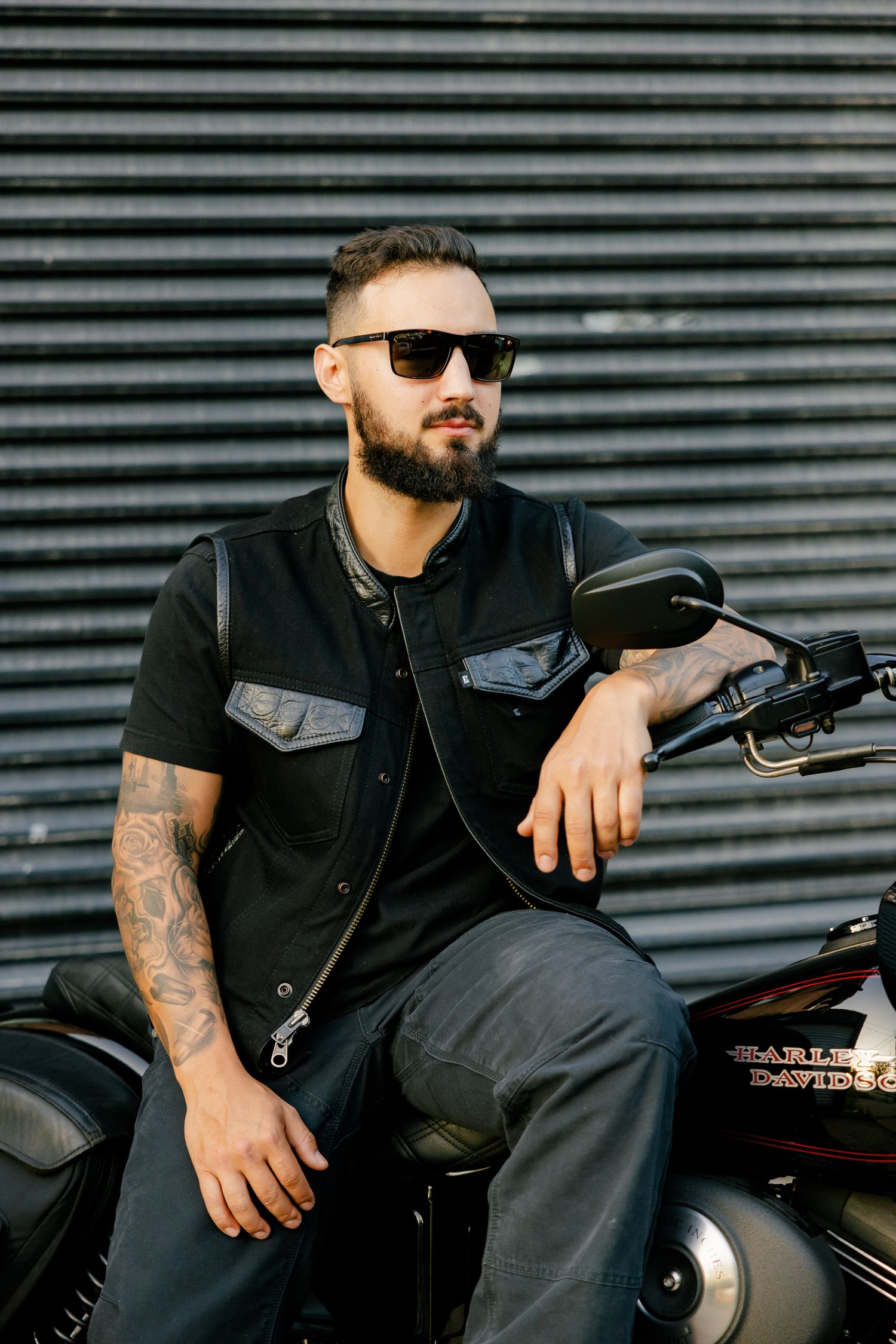 A tattooed rider in dark denim vest and sunglasses leans on their motorcycle handlebars.
