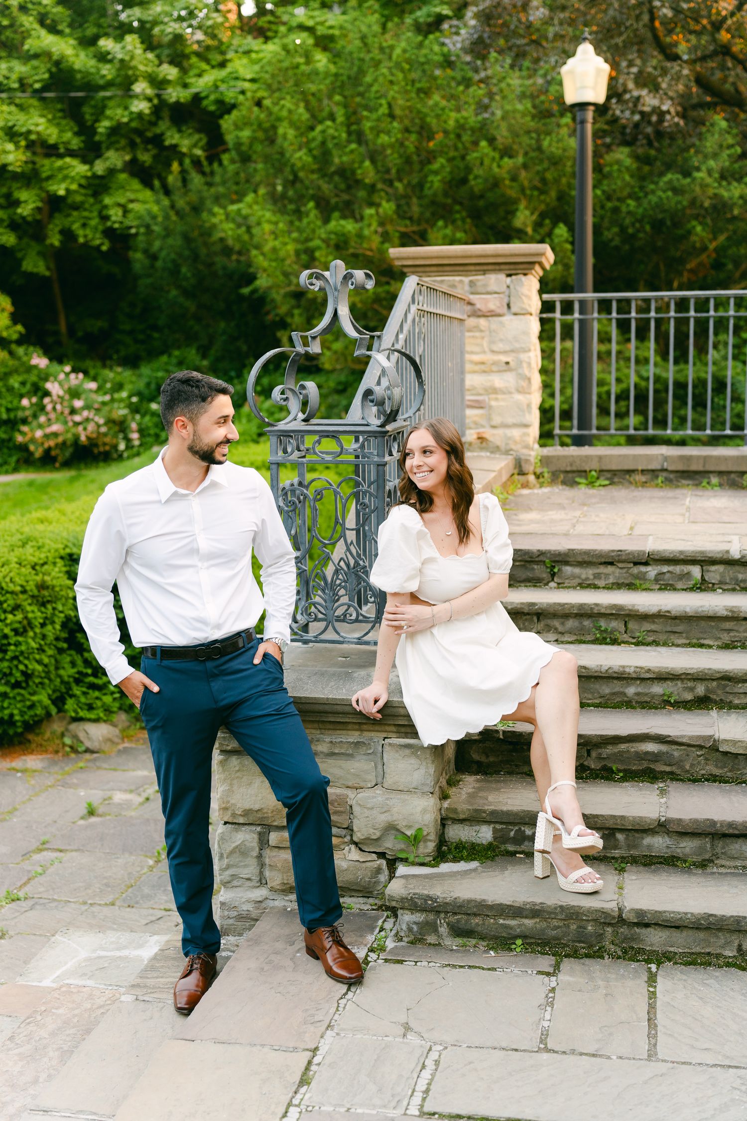 A couple in coordinated white and navy outfits pose elegantly on stone garden steps with ornate railings.
