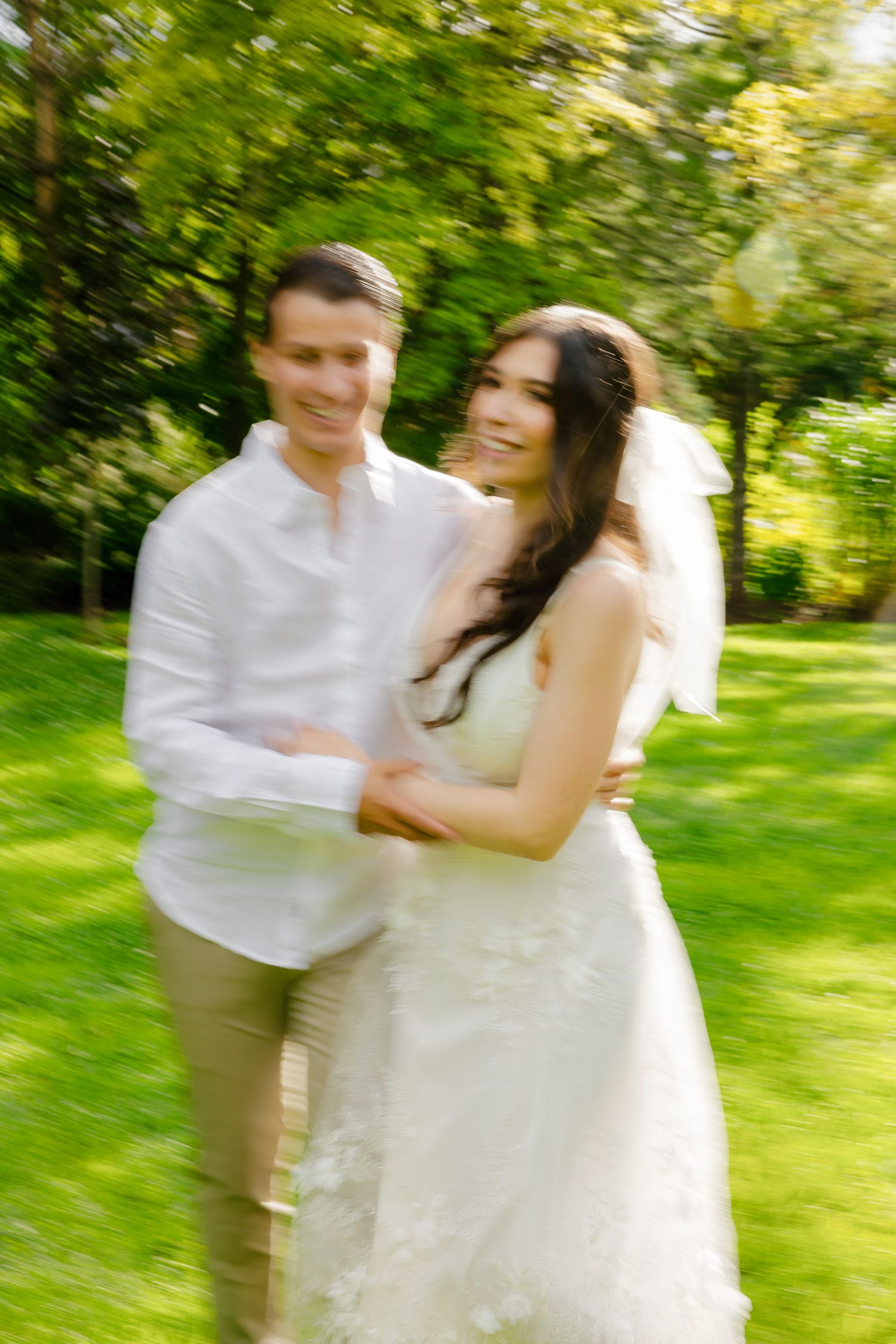 A joyful wedding couple embraces in a sun-dappled garden with vibrant green trees.