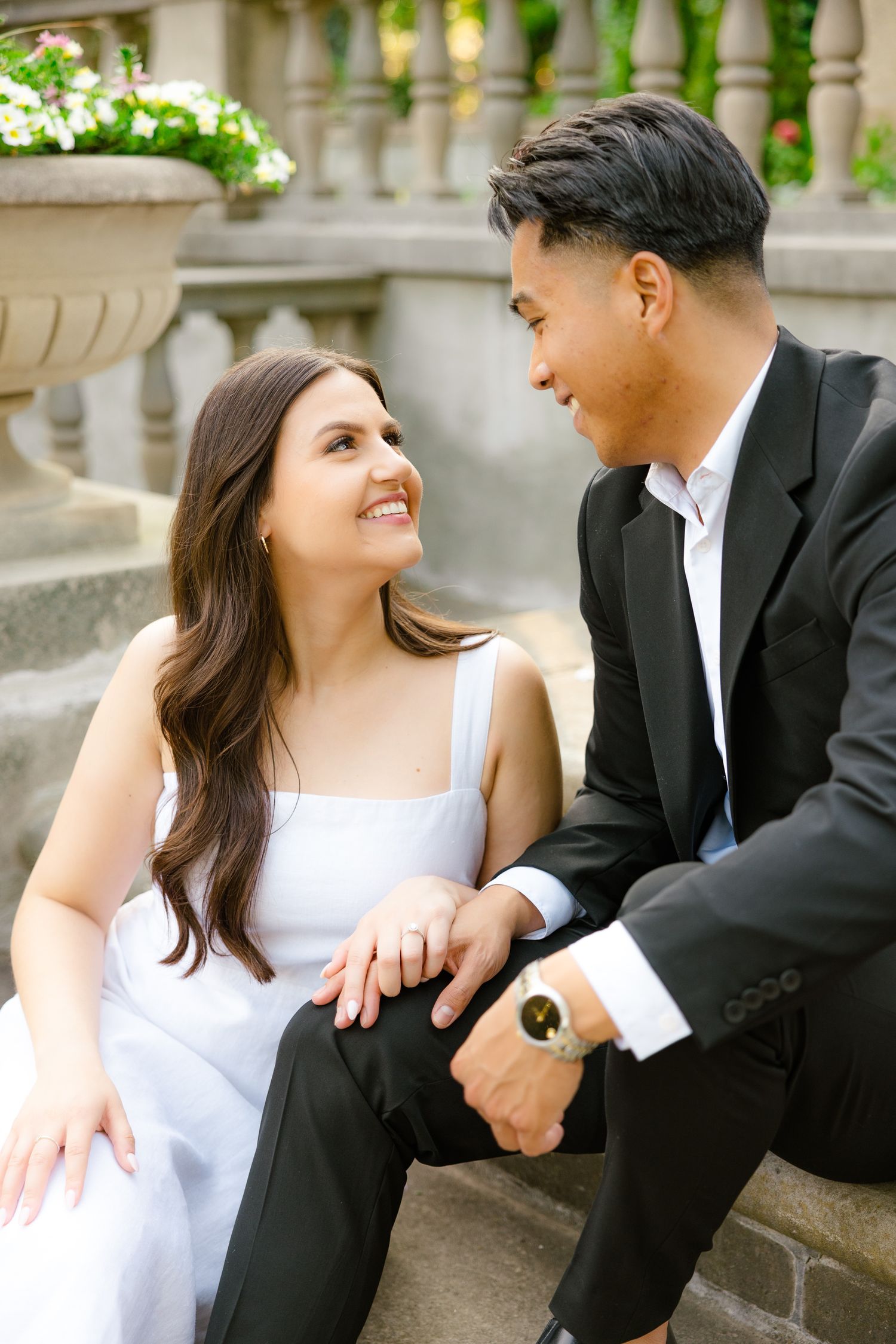 A couple shares joyful expressions while seated on stone steps near decorative garden planters.