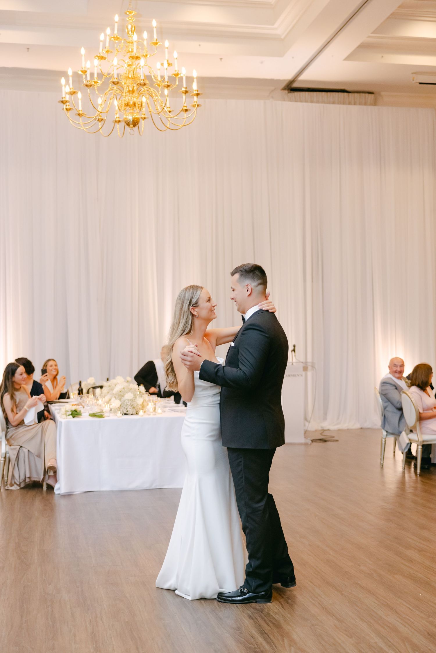 Newlyweds share an intimate dance moment under a gold chandelier in an elegant white reception hall.