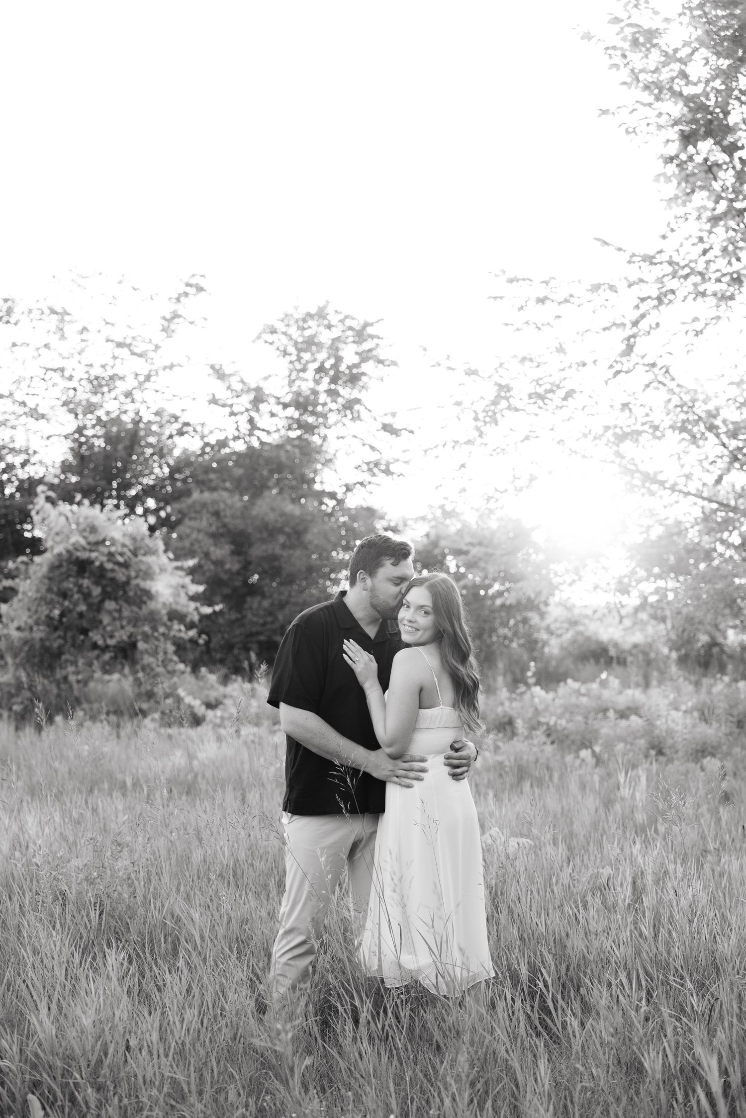 Romantic couple embraces in a sunlit meadow with tall grass and trees at golden hour.