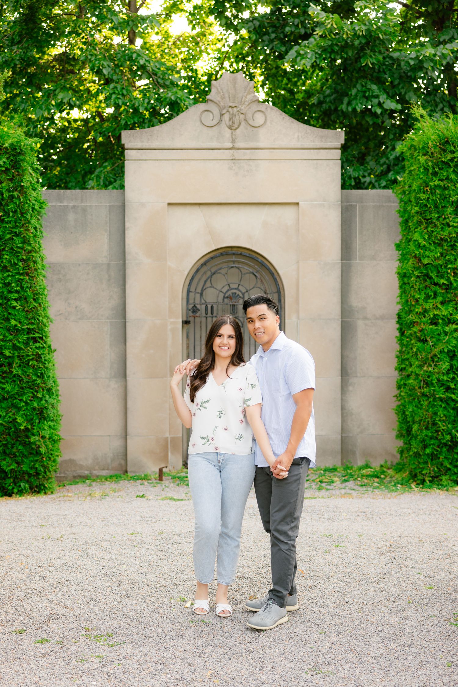 A couple stands together in front of a decorative stone alcove framed by green hedges.