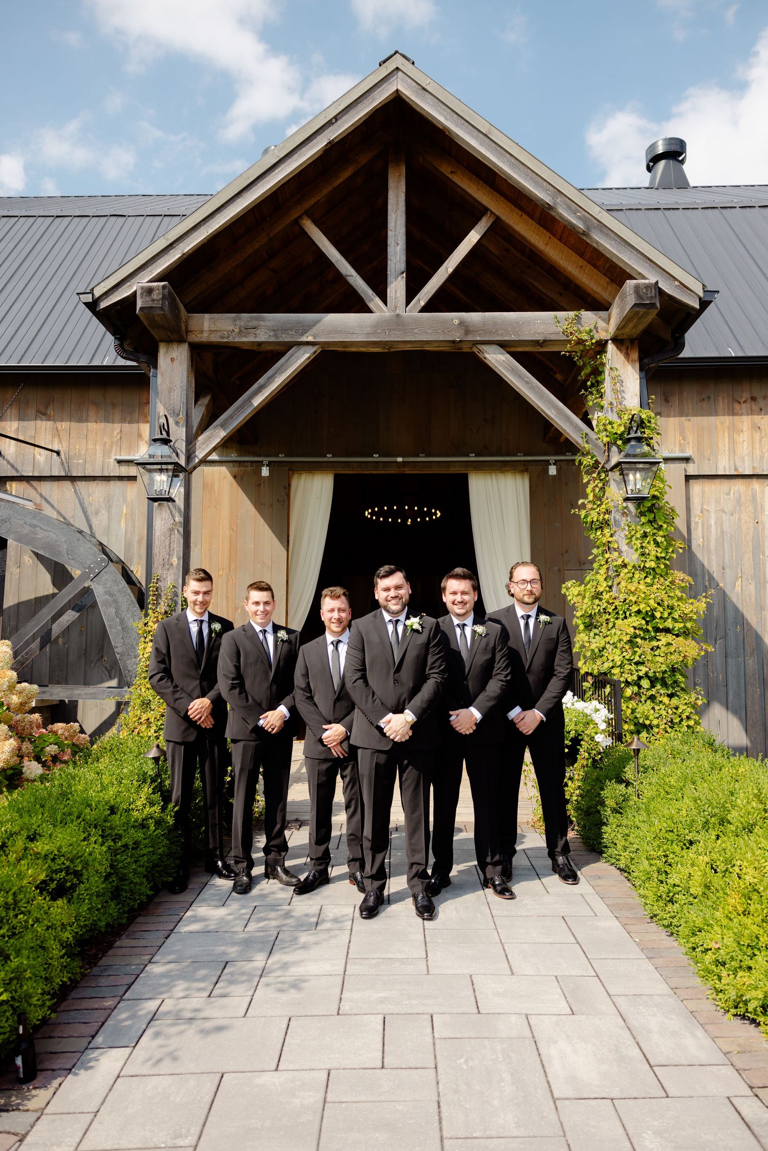 Wedding party in matching dark suits poses formally at rustic barn entrance with stone pathway.