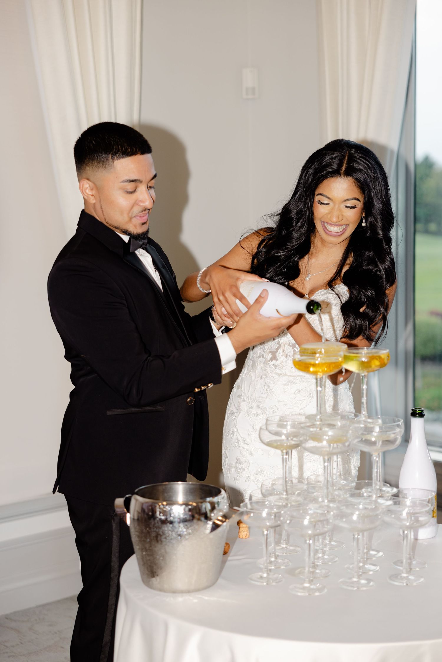 A couple laughs while pouring champagne into a tower of glasses at their wedding reception.