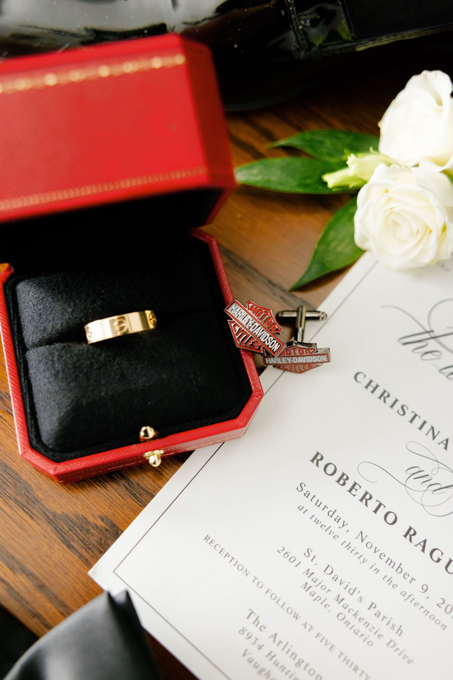 Gold wedding ring in red box next to cufflinks and invitation on wooden surface.