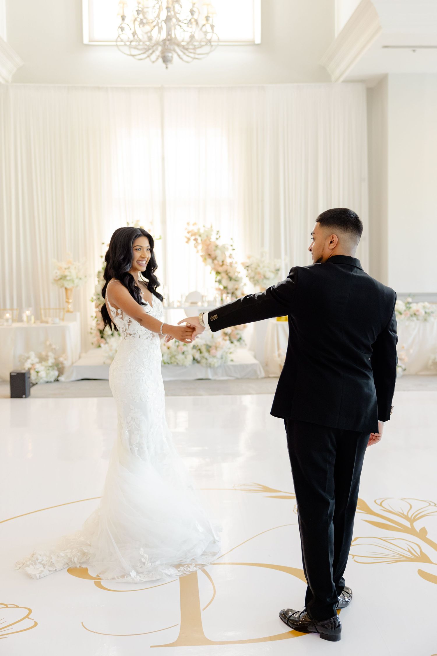 Couple dances together in an elegant white reception venue with ornate chandelier and gold floor details.