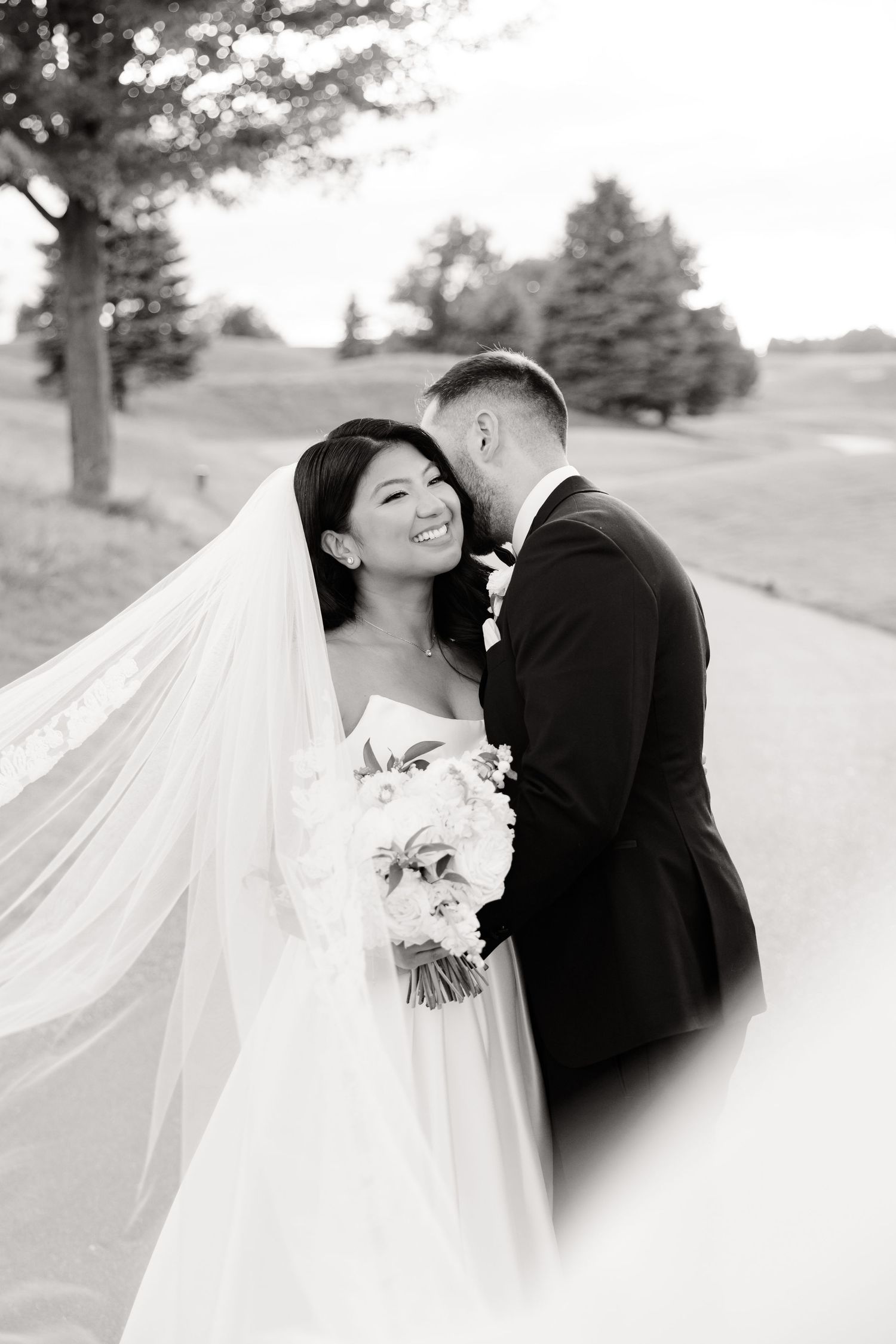 A black and white wedding photo showing a tender moment between newlyweds with flowing veil.
