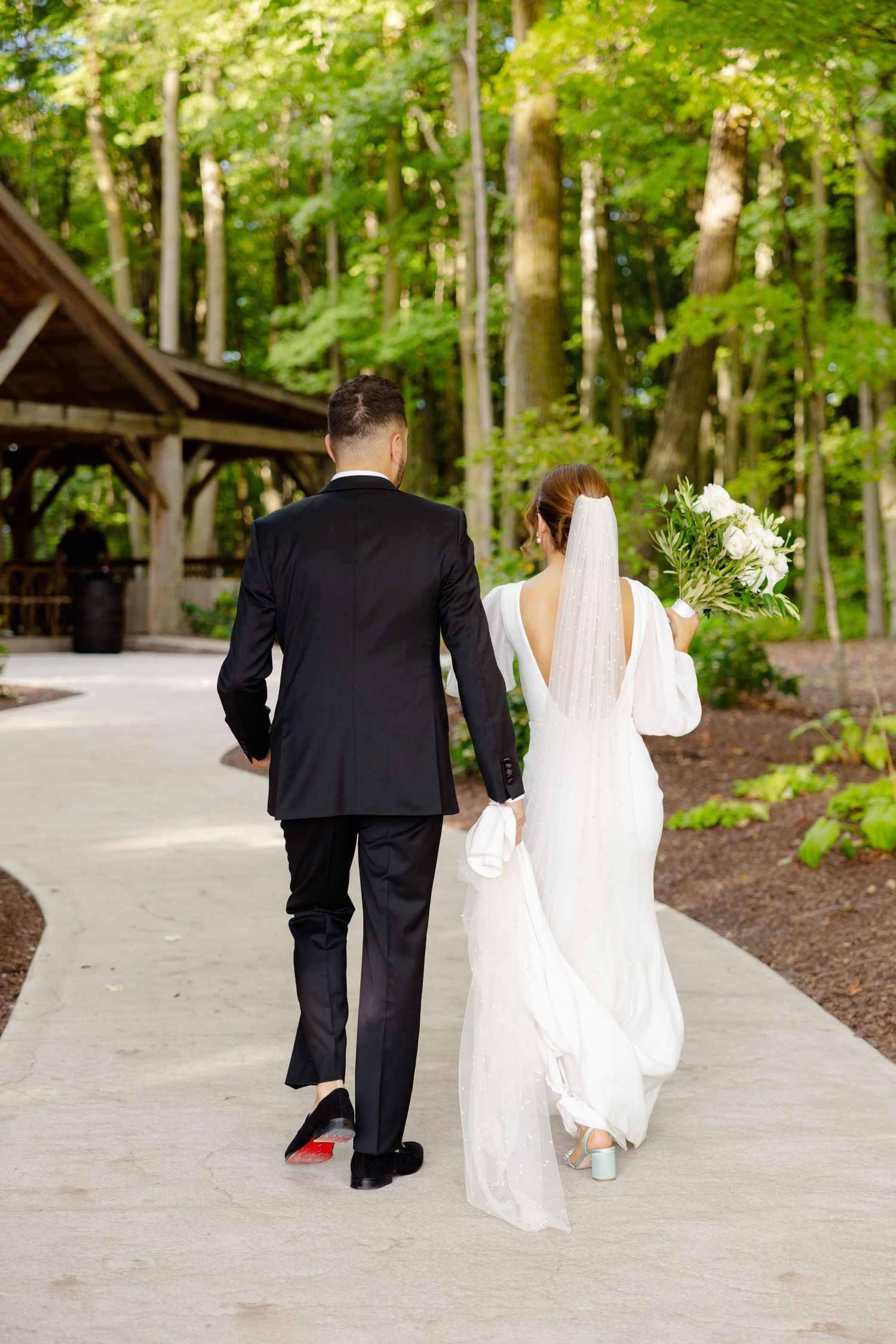 Newlyweds walking away hand in hand on concrete path toward rustic woodland pavilion.