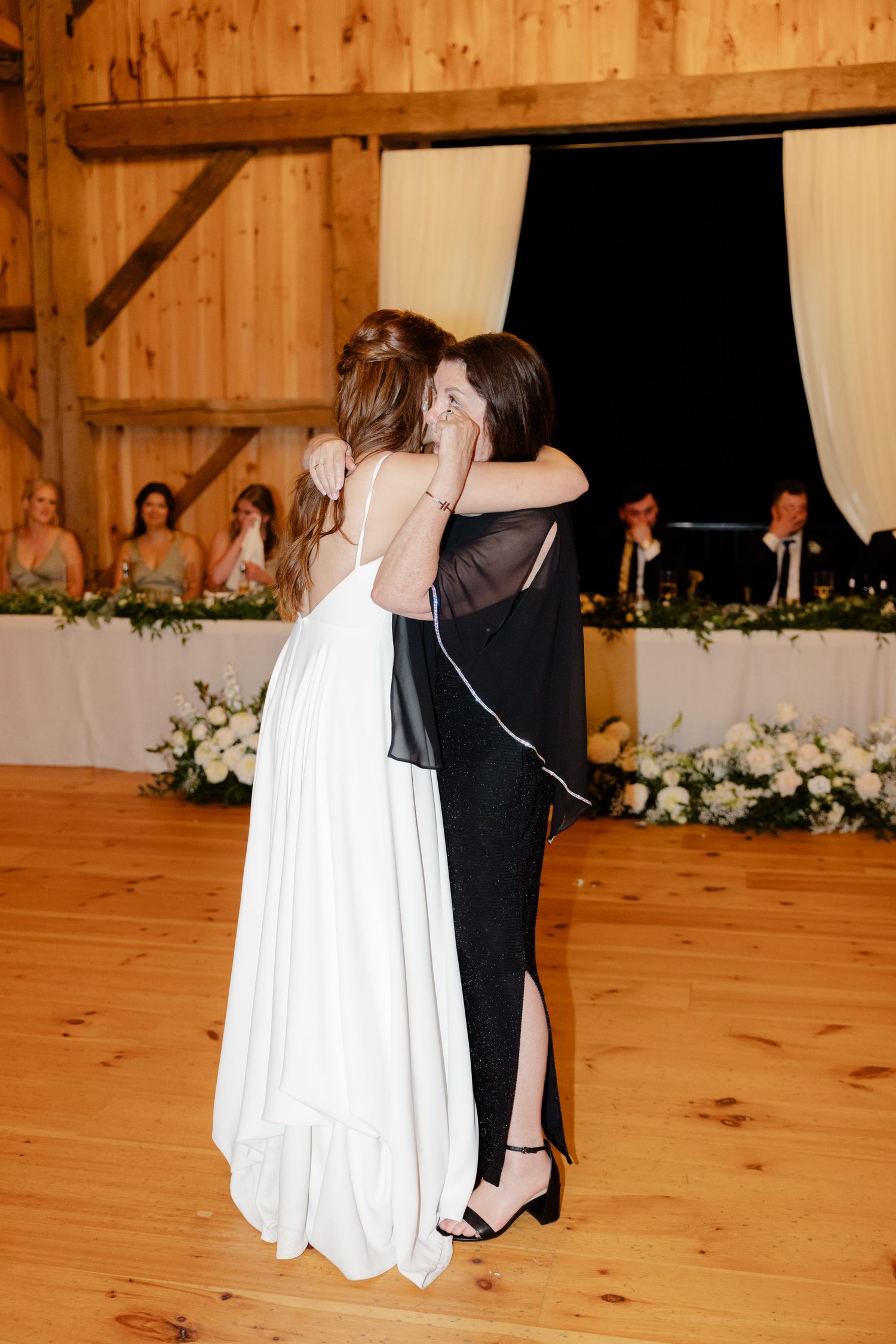 A bride in white hugs a wedding guest wearing black during a rustic barn reception with floral decorations.