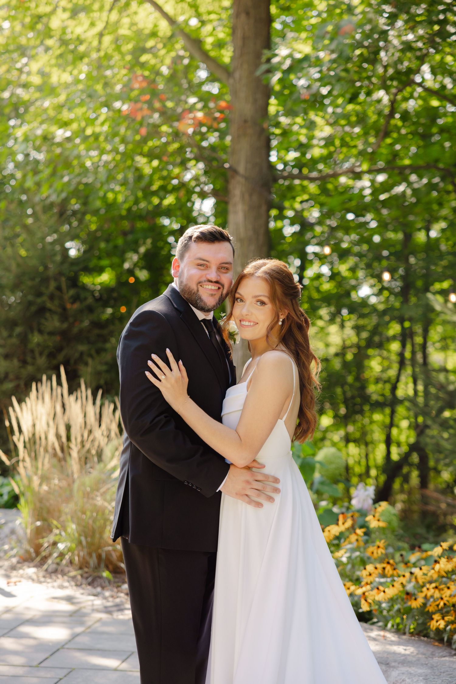 A joyful wedding couple shares an intimate moment on a garden path with sunlit trees in background.