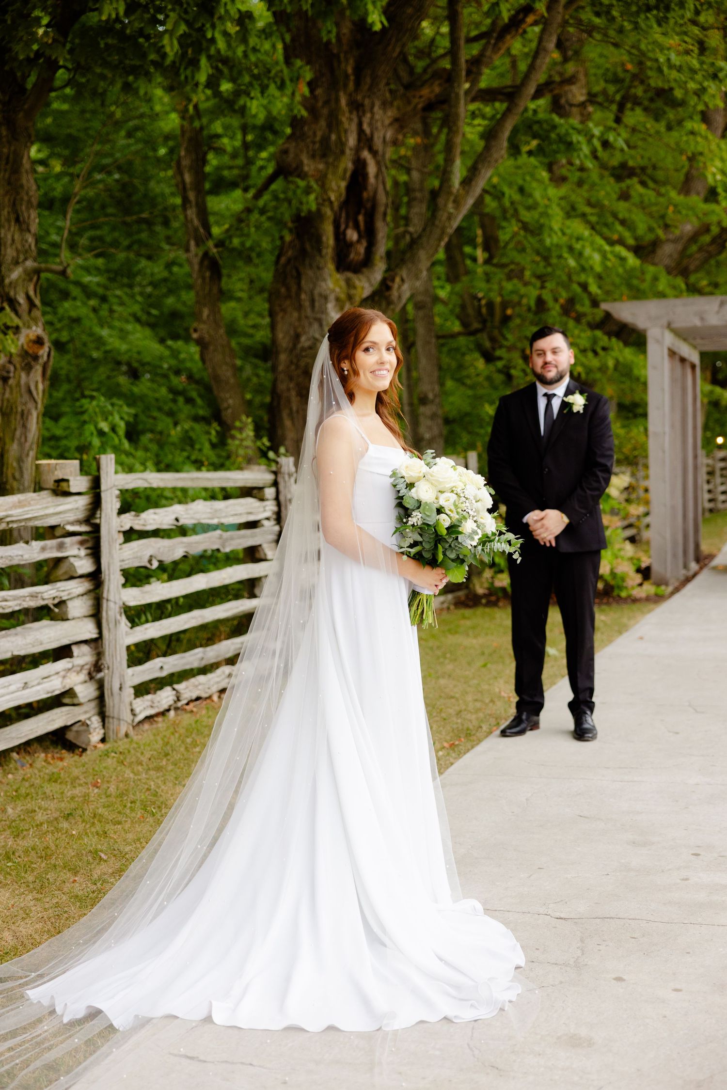 Elegant wedding scene on pathway with white fence and lush greenery backdrop.