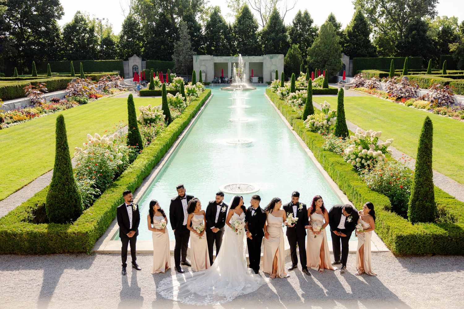 Wedding group photo in front of an elegant garden pool lined with cypress trees and flower beds.
