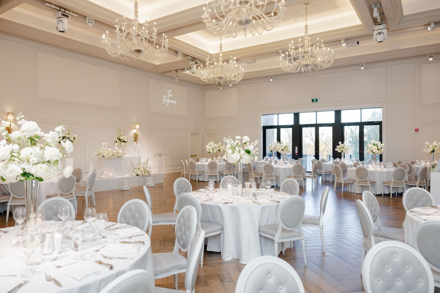 Bright wedding reception venue with crystal chandeliers, white decor, and floor-to-ceiling windows.
