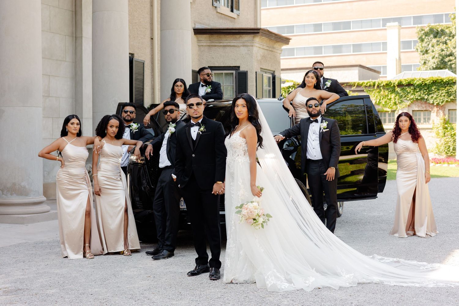 Wedding party in formal attire poses together outside a columned building with a black luxury SUV.