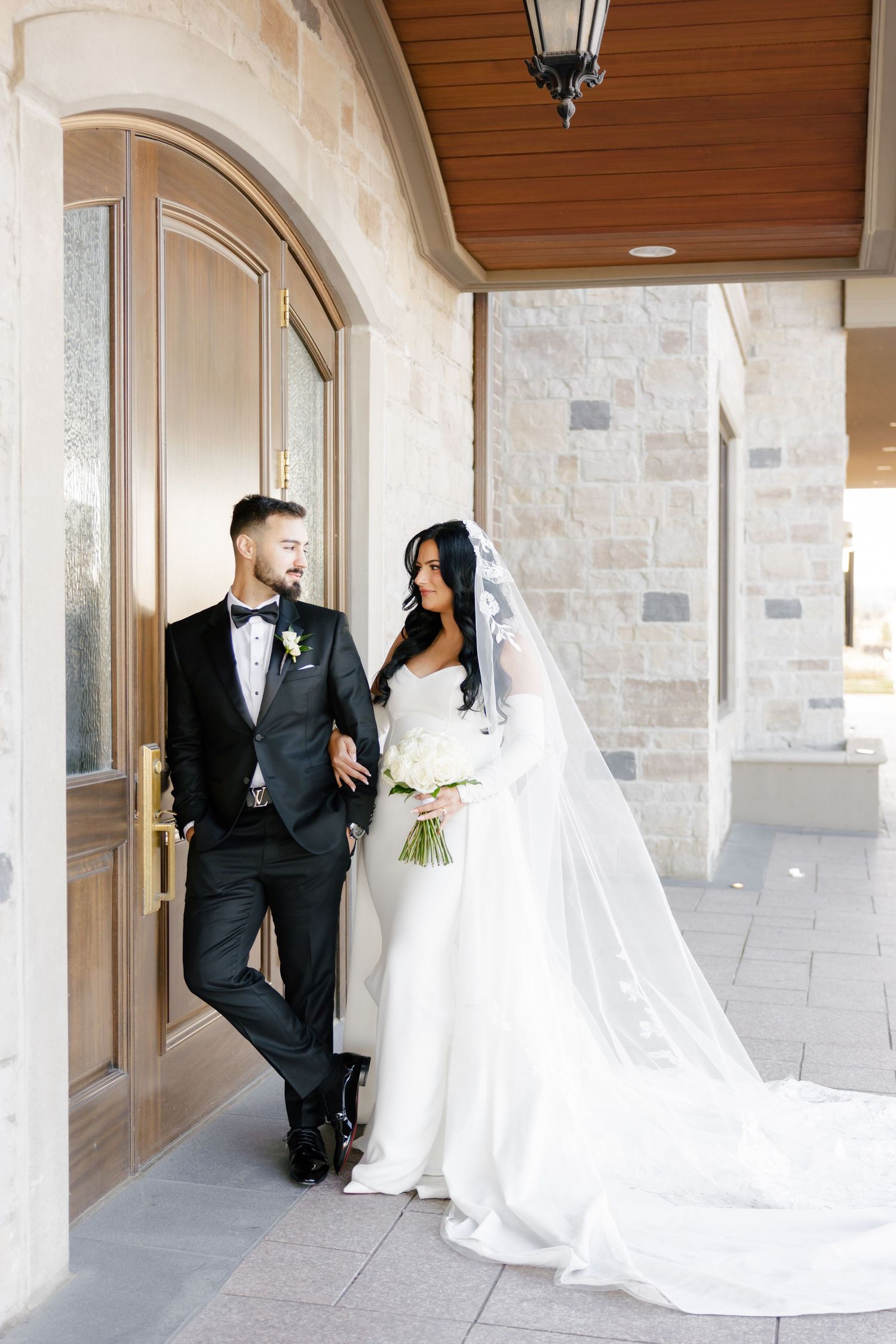 Bride and groom stand beneath arched entrance with wooden ceiling detail.