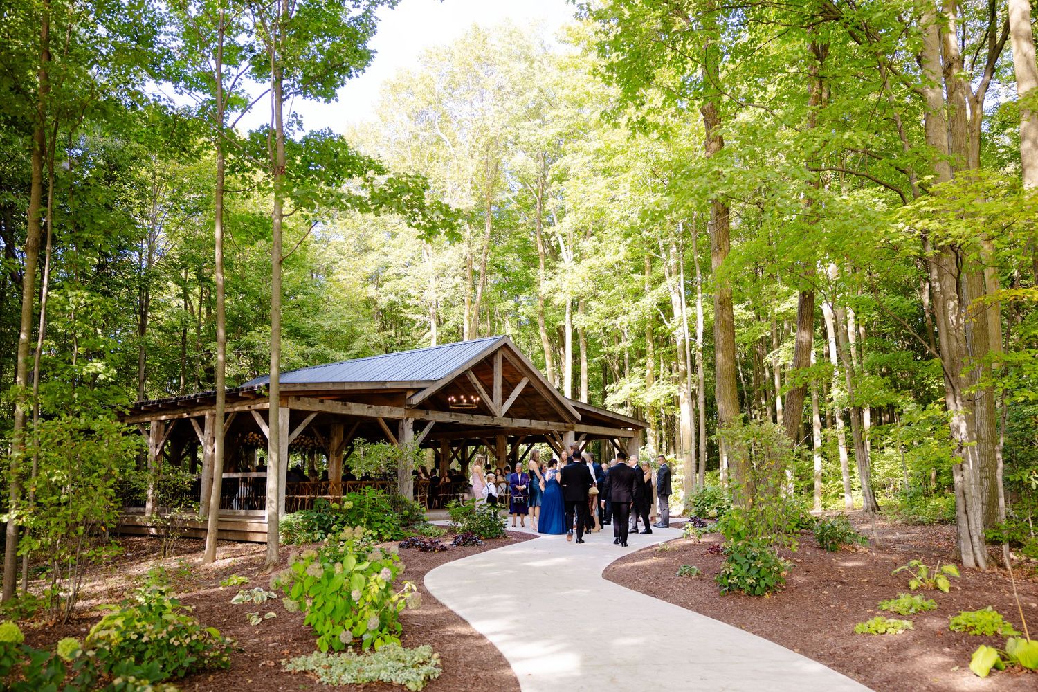 Rustic wooden pavilion venue nestled in lush forest setting with paved walkway approach.
