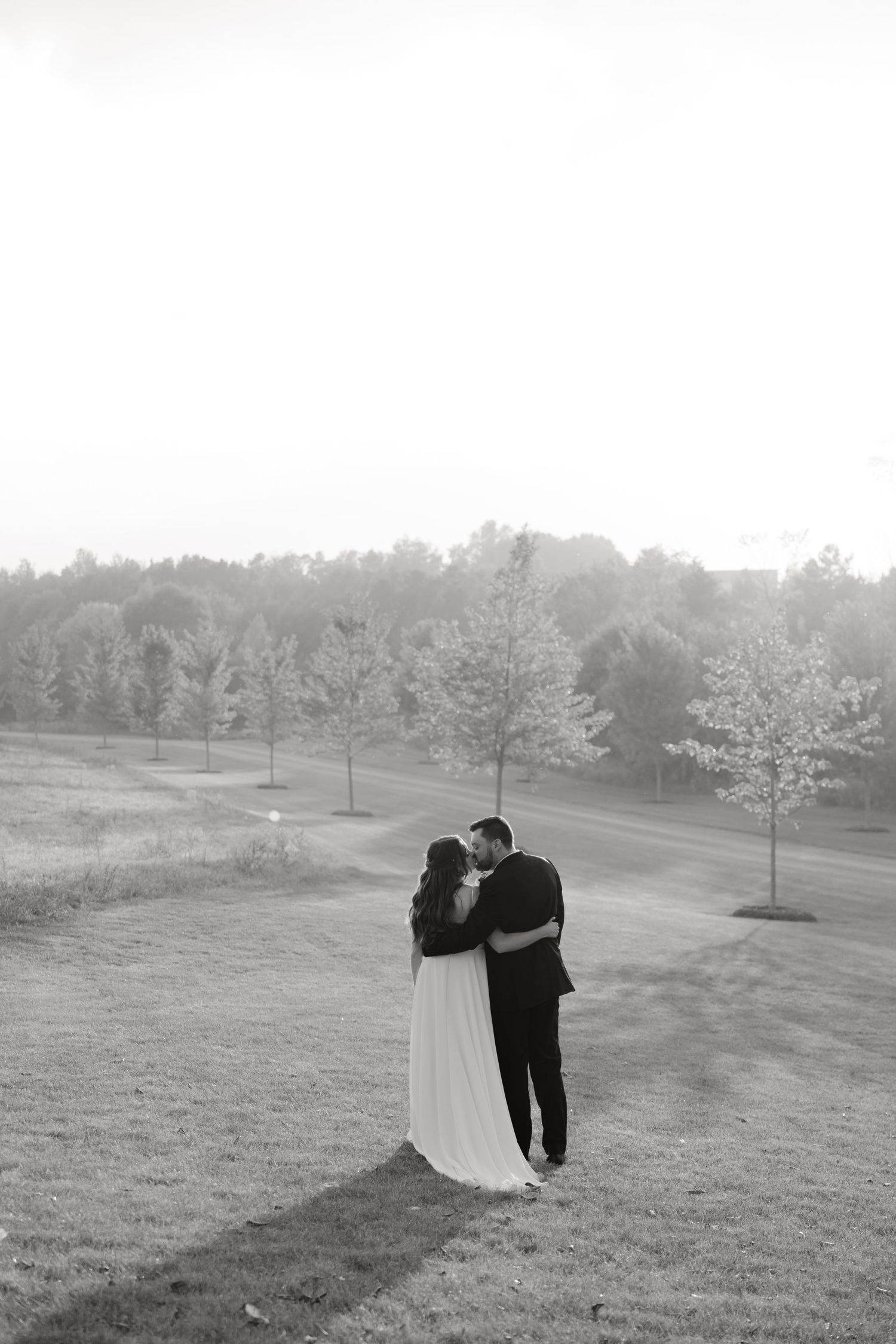 Black and white wedding portrait of a couple embracing on a hillside at sunset.