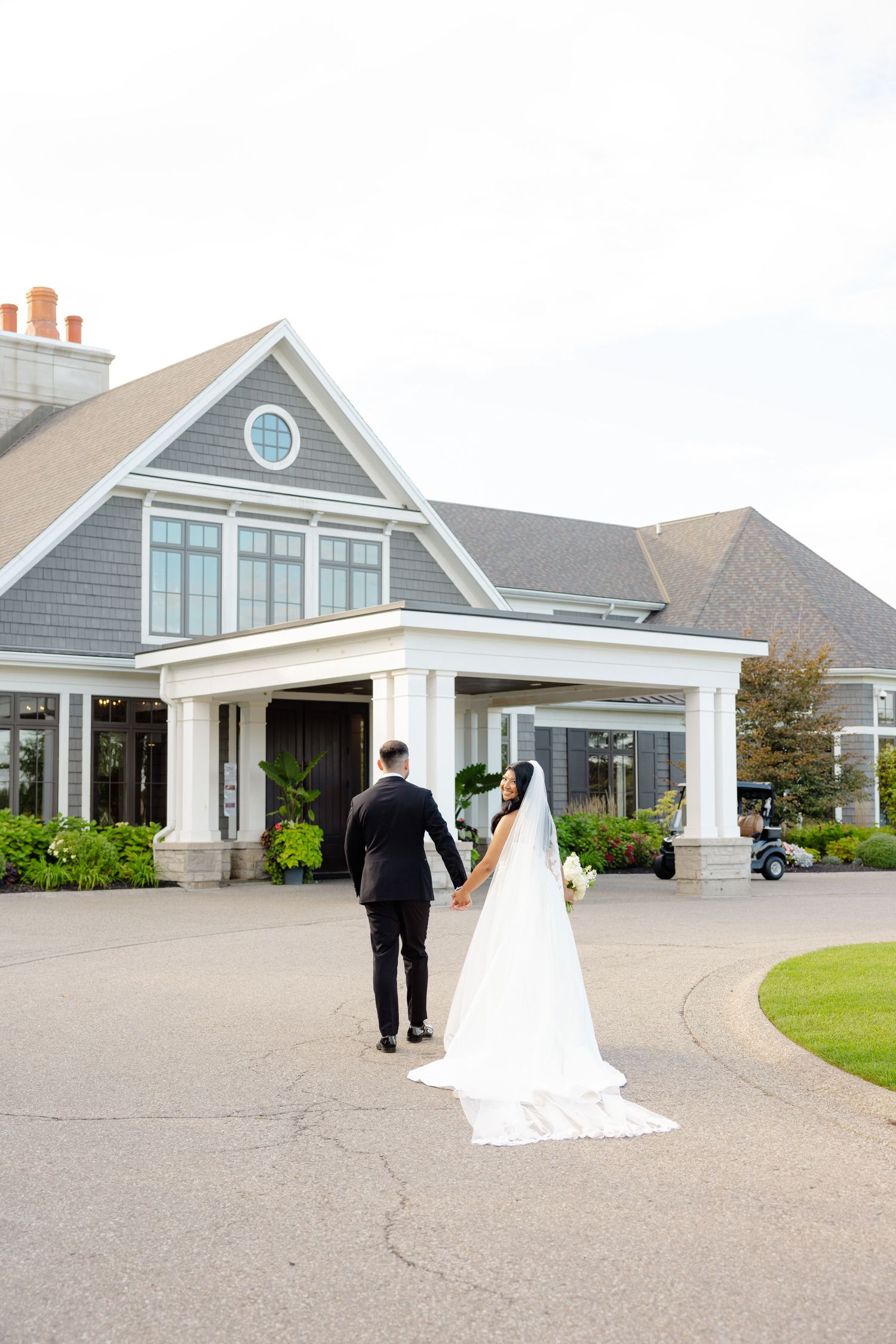 A couple in wedding attire walks up to an elegant gray house with white trim and columns.