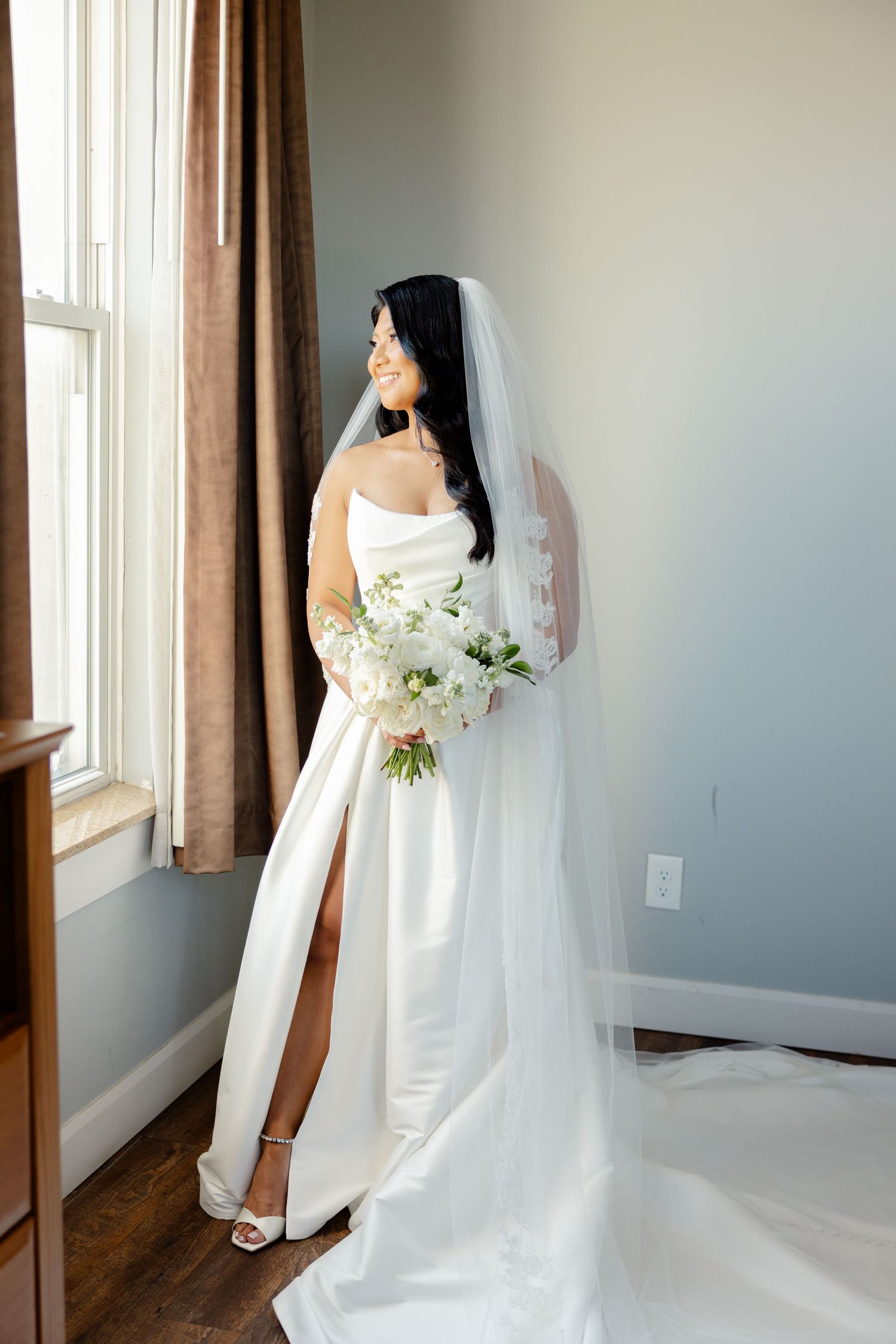 Elegant bride in white dress with thigh slit gazes out window while holding classic white bouquet.