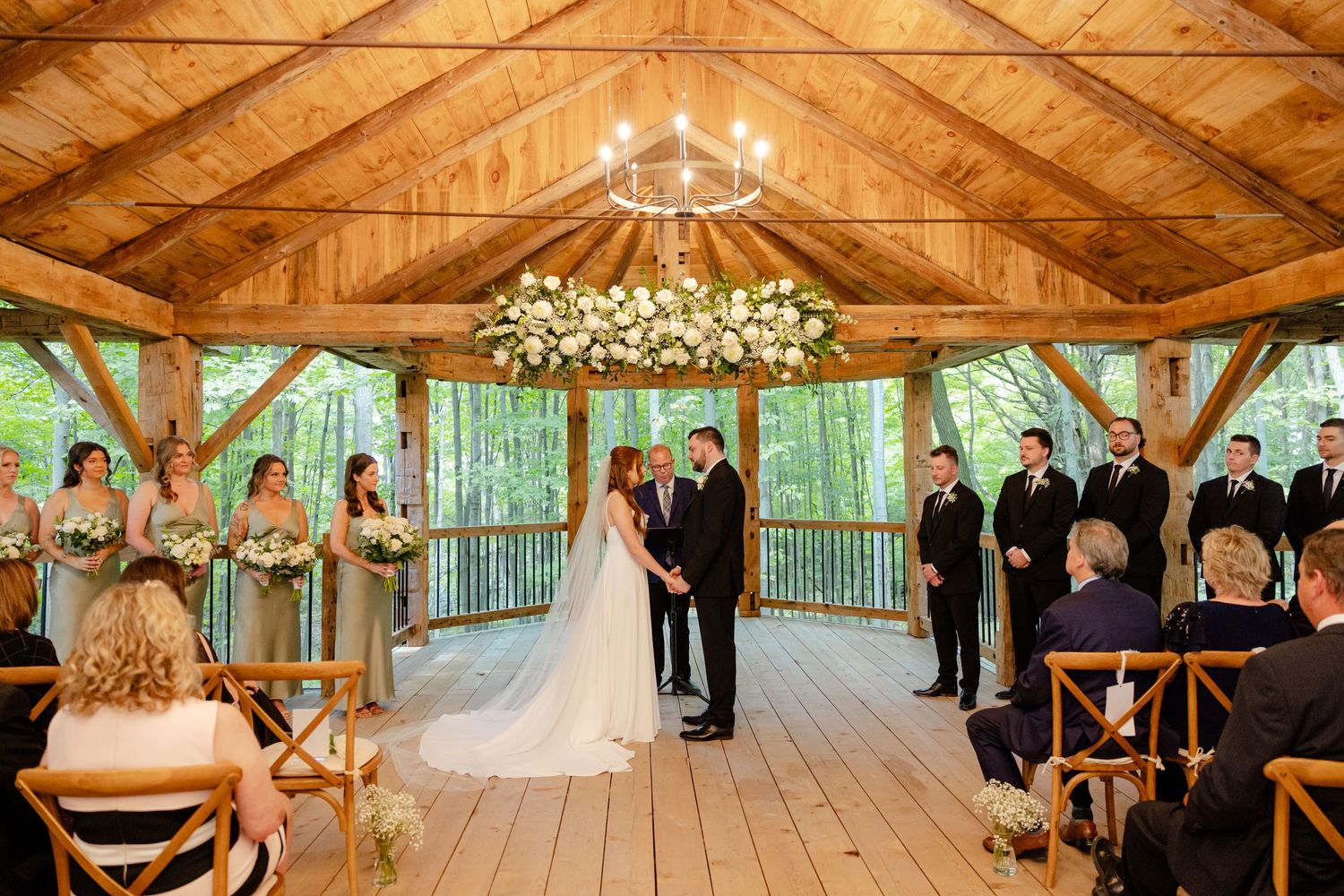 Wide view of wedding ceremony in rustic wooden pavilion surrounded by lush forest.