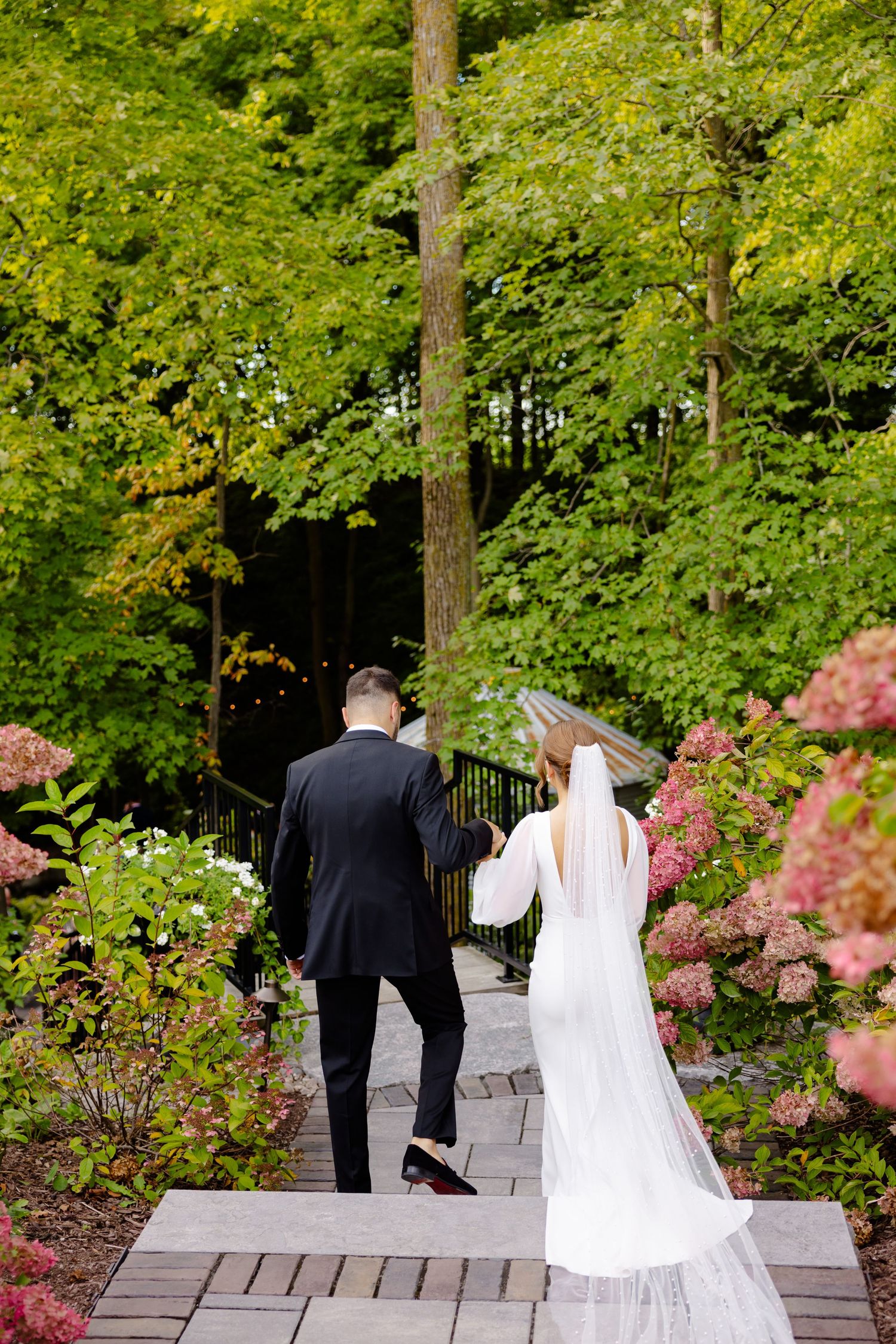 Wedding couple walking up garden steps surrounded by blooming azaleas and trees.
