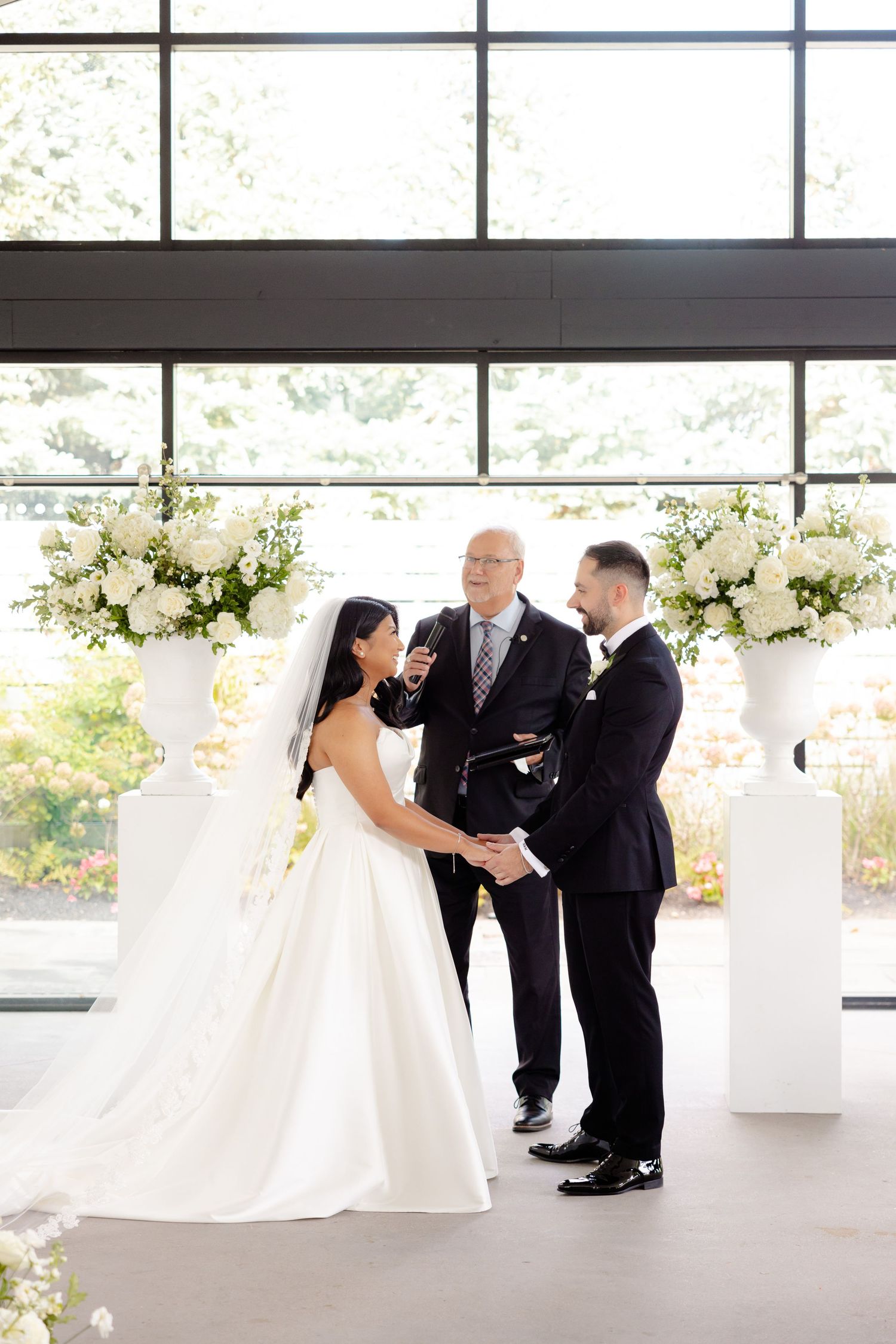 Wedding ceremony at an elegant venue with white floral pillars and large windows.