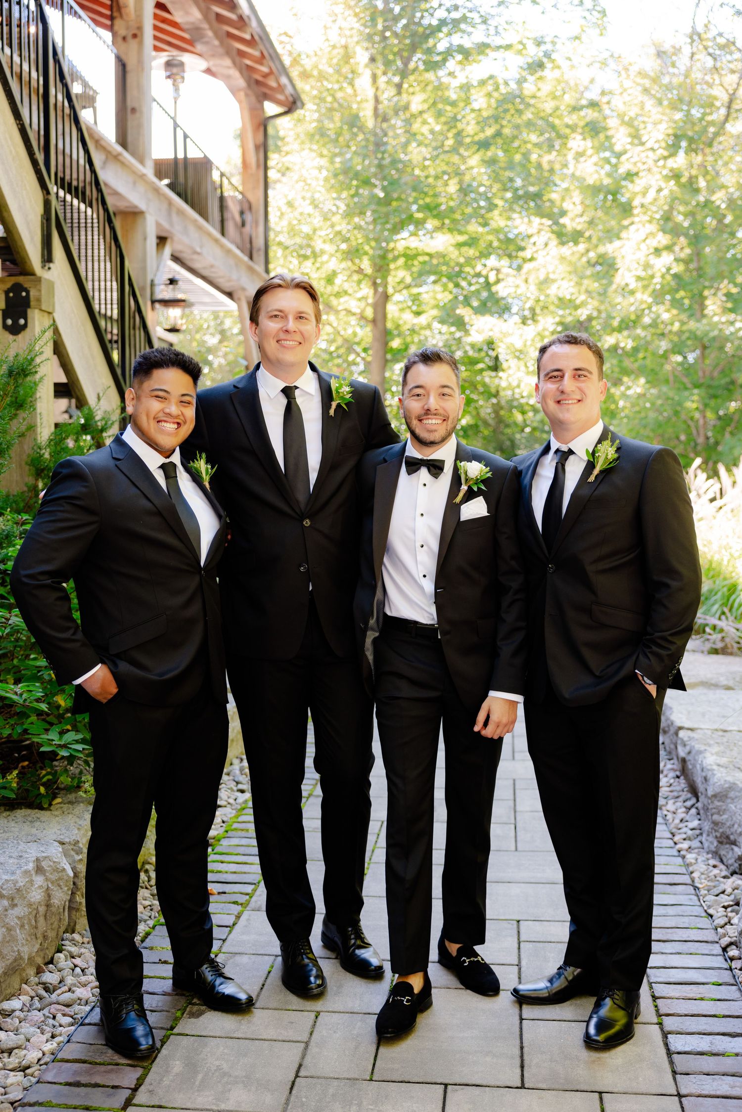 Groomsmen in dark suits pose casually outside venue with stone pathway and greenery.