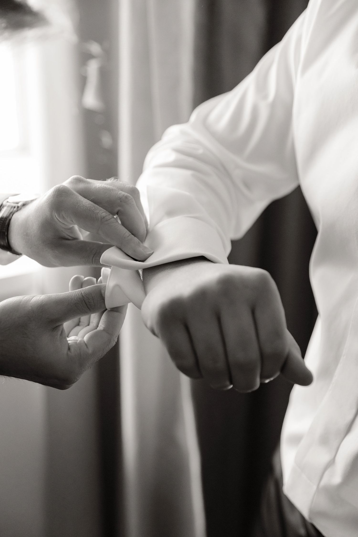 A black and white close-up photograph of hands adjusting cufflinks on a white dress shirt sleeve.
