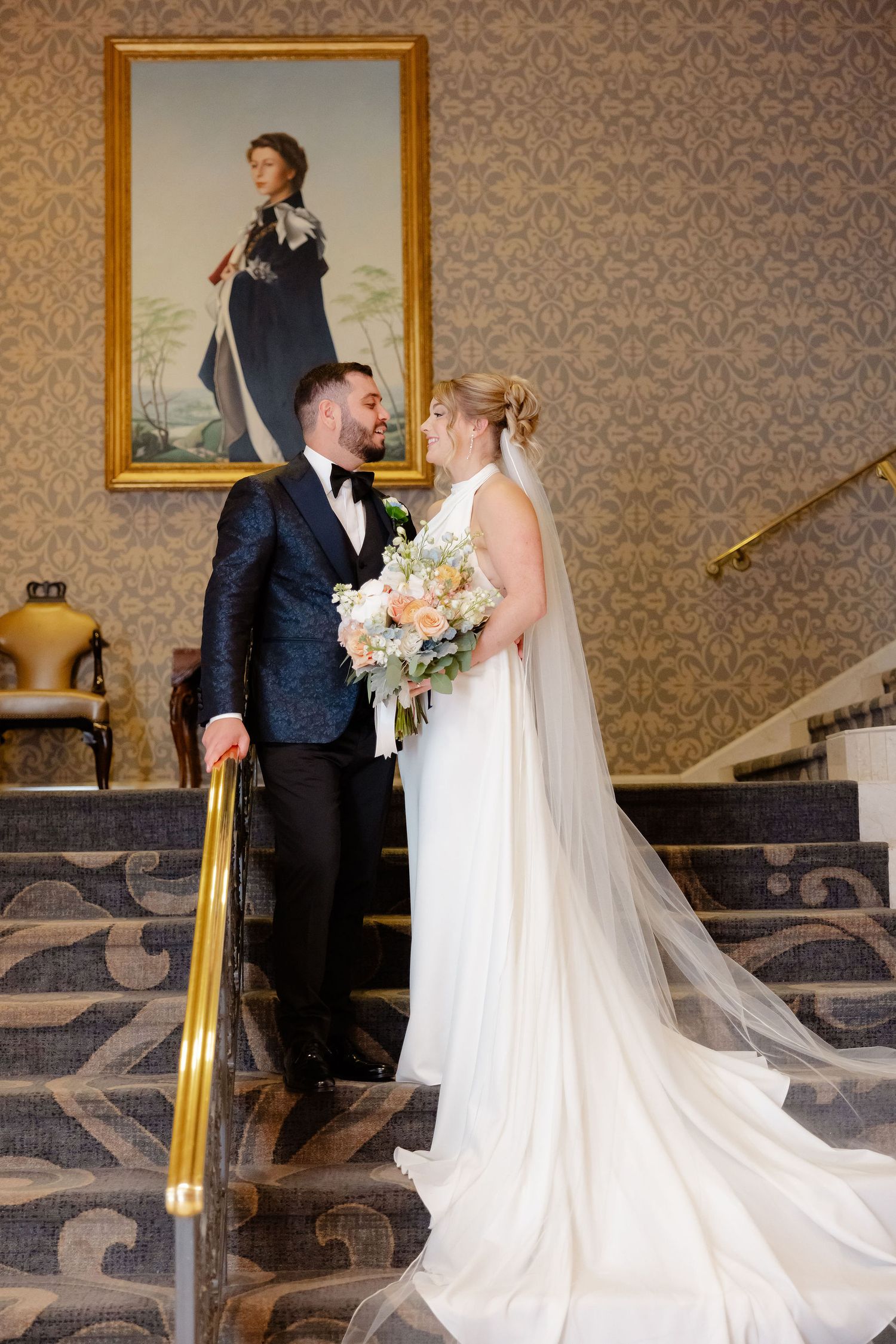 A romantic wedding couple shares an intimate moment on a grand staircase with ornate wallpaper and gold frames.