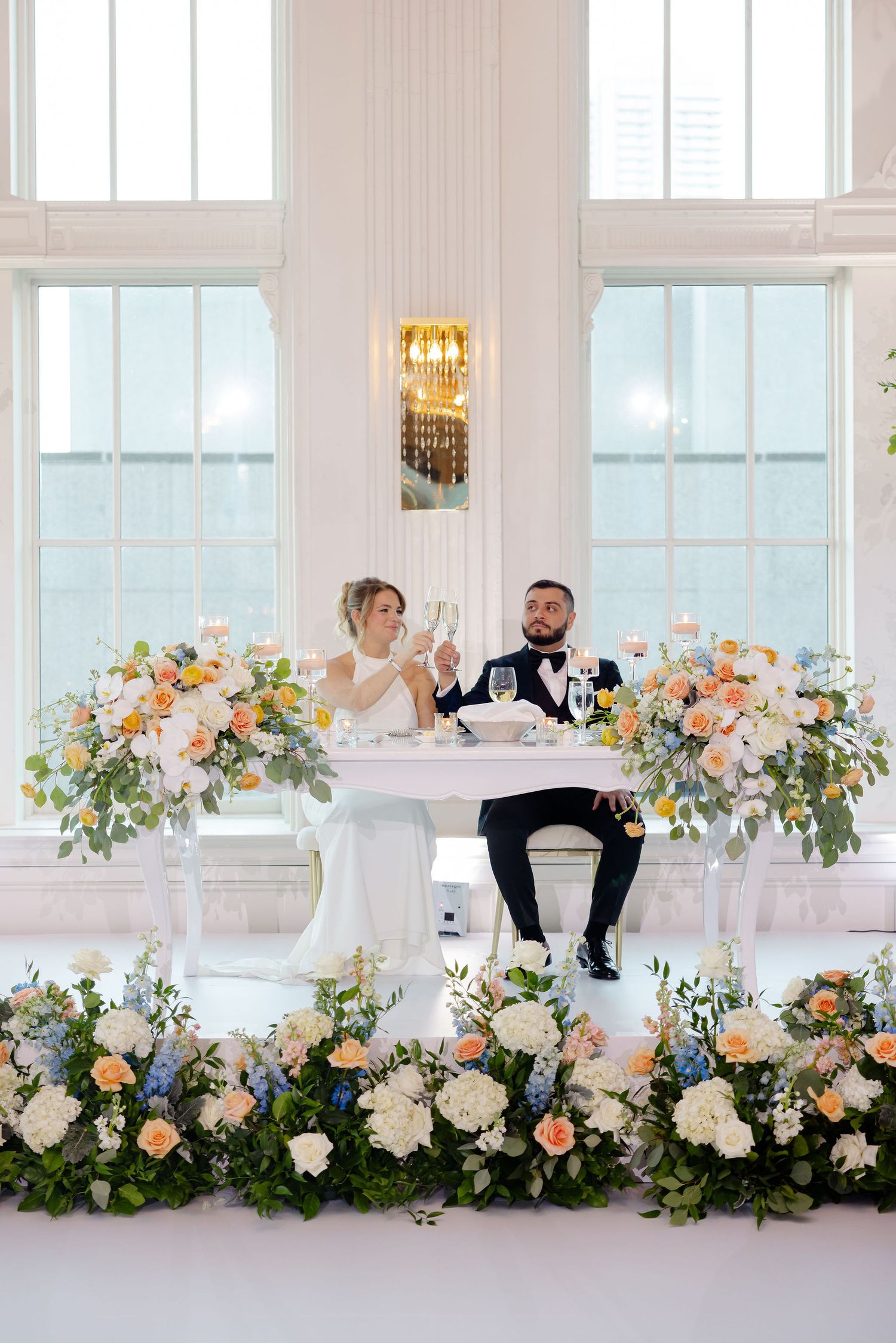 Wedding couple at white reception venue with luxurious floral table decorations in peach and cream tones.