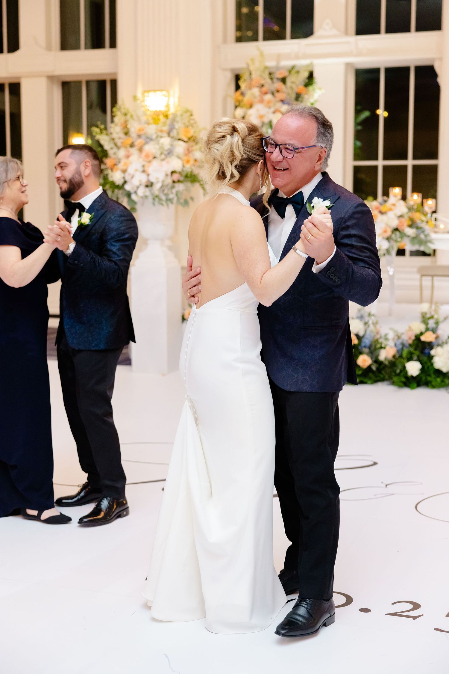 Newlyweds dancing together in an elegant white ballroom decorated with floral arrangements.
