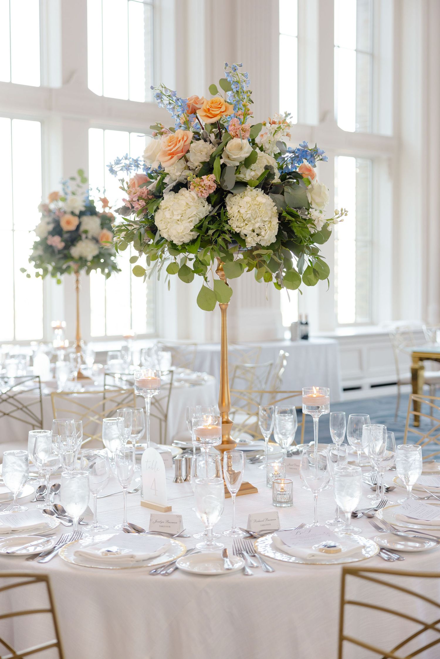Luxurious wedding reception tables with tall floral centerpieces, white linens, and crystal glassware in a bright venue.